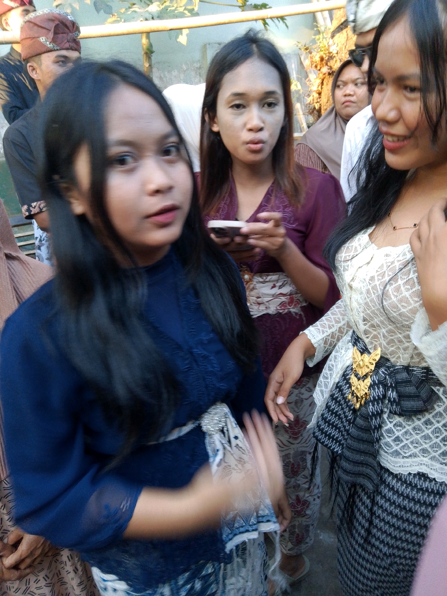 The image shows a group of people, likely at an outdoor event in Kuripan Utara, Indonesia. The foreground is dominated by two young women. The woman on the left, facing slightly away from the camera, wears a dark blue traditional outfit with embroidered details. Her dark hair is long and frames her face. She is holding her left arm out, with her hand blurred by motion. The woman on the right is in profile, smiling and looking towards her right. She wears a white lace top with intricate patterns and a dark sarong with a checkered pattern. A prominent gold brooch is attached to her waist. Behind these two women, other individuals are visible, including a woman wearing a maroon headscarf and a woman in a maroon top. A man wearing a traditional red and patterned headdress is also partially visible in the background, along with other people and foliage. The environment appears to be a gathering with a simple backdrop suggesting an outdoor setting.