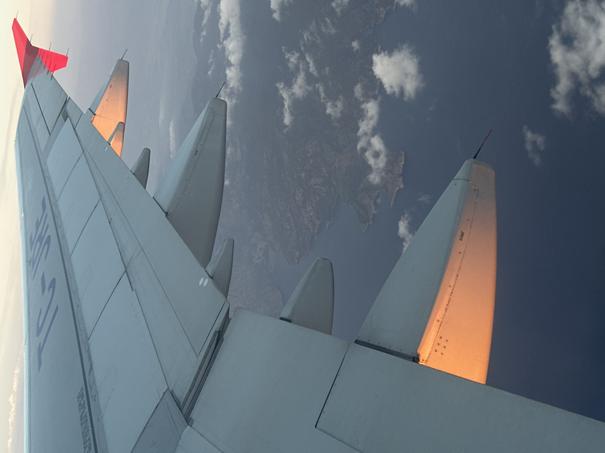 An aerial photograph captures the starboard wing of an aircraft in flight, with the registration "TC-JRE" visible on its upper surface, indicating Turkish registry. The wing, predominantly light grey, features multiple flap segments along its trailing edge and a red wingtip fence at its extremity. A strong, low-angle sunlight, indicative of either sunrise or sunset, casts an intense golden-orange glow on the underside of a main flap and a lower wing section, contrasting with the overall muted tones of the aircraft and the environment.
Below the aircraft, a vast expanse of dark blue water is visible, dotted with numerous irregular, rocky landmasses consistent with islands or a highly indented coastline. Scattered white cumulus clouds are present both above and below the wing, casting diffuse shadows on the water and land. The sky above the wing is a hazy light blue-grey. No individuals are depicted. The geographical features, while characteristic of coastal Türkiye, do not permit identification of a specific city within Türkiye.