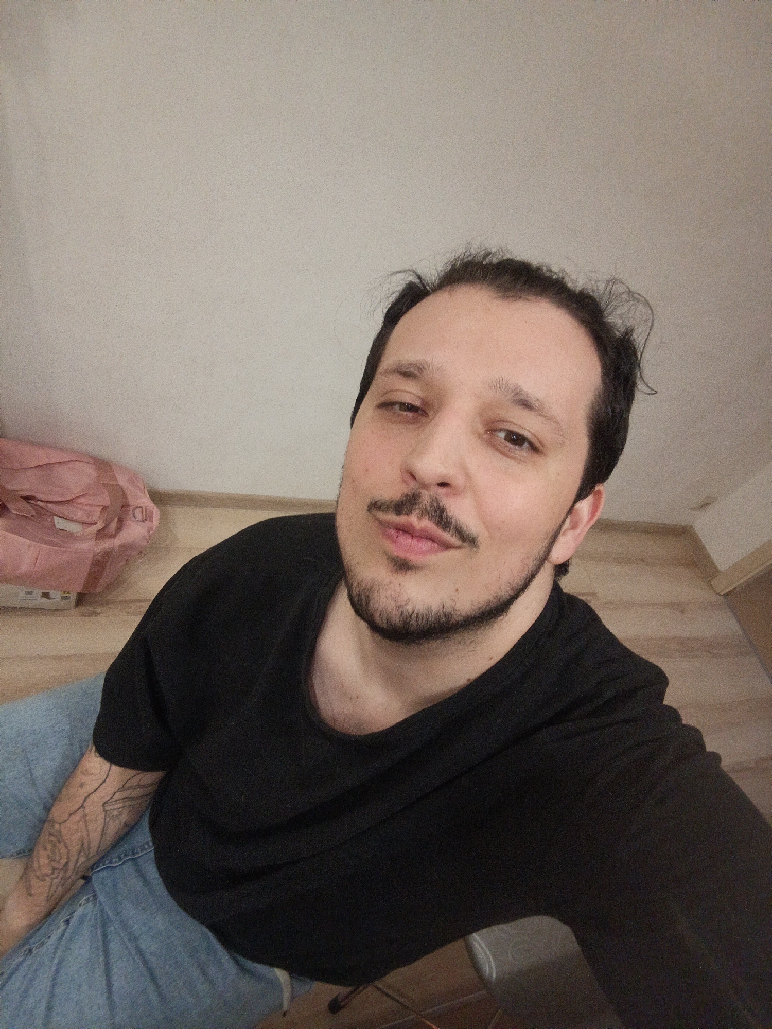 A man with dark hair, a mustache, and a beard is positioned in the foreground, wearing a black t-shirt and blue jeans. Part of a tattoo is visible on his left arm. Behind him is a cream-colored wall above wooden-patterned flooring. A pink object, possibly a bag, and a box are visible in the background, against the wall. There is no specific evidence in the image to determine the location in the city of Penza, Russia.