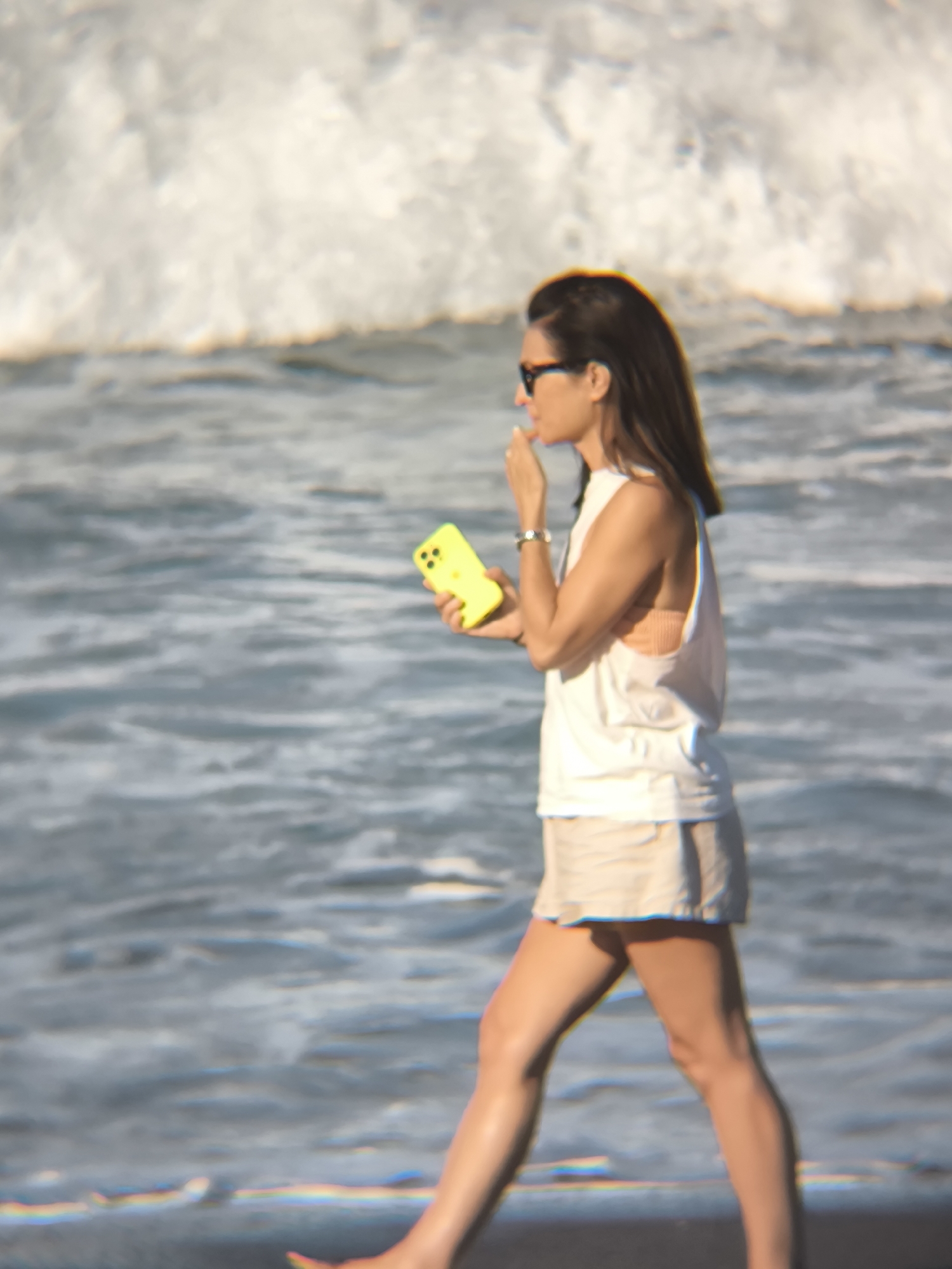 A woman walks barefoot along the beach in North Kuta, Indonesia, holding a bright yellow cell phone and touching her lips. She is wearing sunglasses, a white tank top, beige shorts, and a watch on her left wrist. The beach stretches along the foreground, with water and a breaking wave in the background.