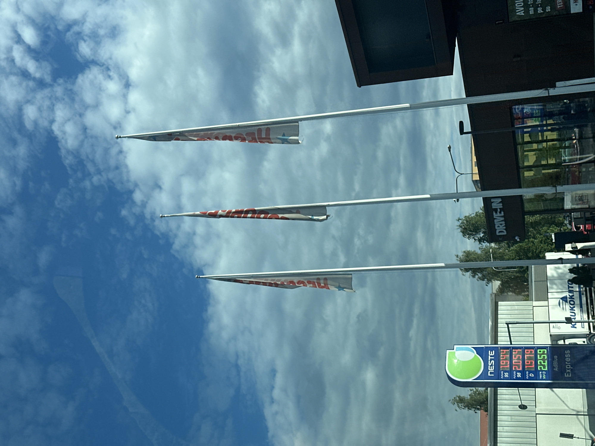 This is a medium shot of a gas station under a cloudy blue sky. The gas price sign displays prices for "NESTE FUTURA 95", "NESTE FUTURA 98", "NESTE D", and "NESTE MYD". Three tall flags are planted in front of a building with a sign that reads "DRIVE-IN". A white truck with "KAUKOKITO" written on its side is parked near the building. The overall scene suggests a commercial area with services like a gas station and a drive-in facility.