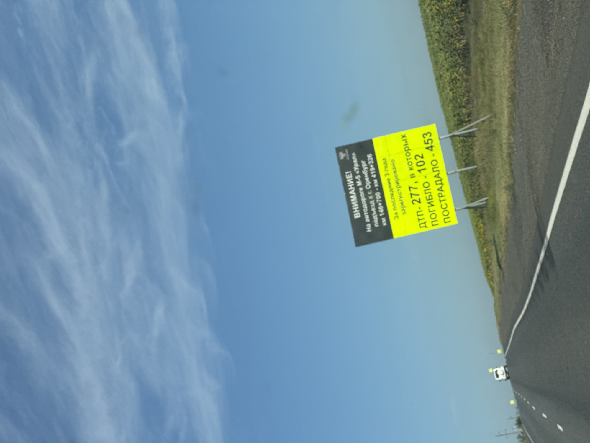 A road sign with a yellow background and black text is visible in the center of the image. The sign reads "ВНИМАНИЕ!" (ATTENTION!) and provides statistics about traffic accidents, including the number of deaths and injuries. The sign is positioned alongside a road in what appears to be a rural or semi-rural area, with a field of sunflowers visible to the right. The sky is blue with some wispy clouds. A white car is driving away from the camera on the road. The image is taken from a moving vehicle, as indicated by the slight motion blur. There is no visible indication that this location is the city of Burdygino, Russia.