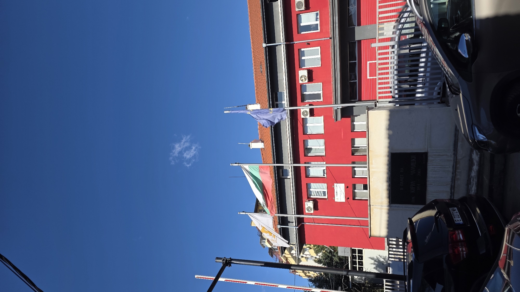 The image depicts a street scene in Blagoevgrad, Bulgaria. Dominating the view is a red building, possibly a public or governmental institution, adorned with three flags: the European Union flag, the Bulgarian national flag, and the flag of Blagoevgrad. The building features multiple windows, some equipped with air conditioning units, and a commemorative plaque is visible on its facade. A barrier arm with red and white stripes is present, suggesting a controlled access point. Two cars are partially visible on the right side of the frame. The sky is clear and blue, with a small white cloud.