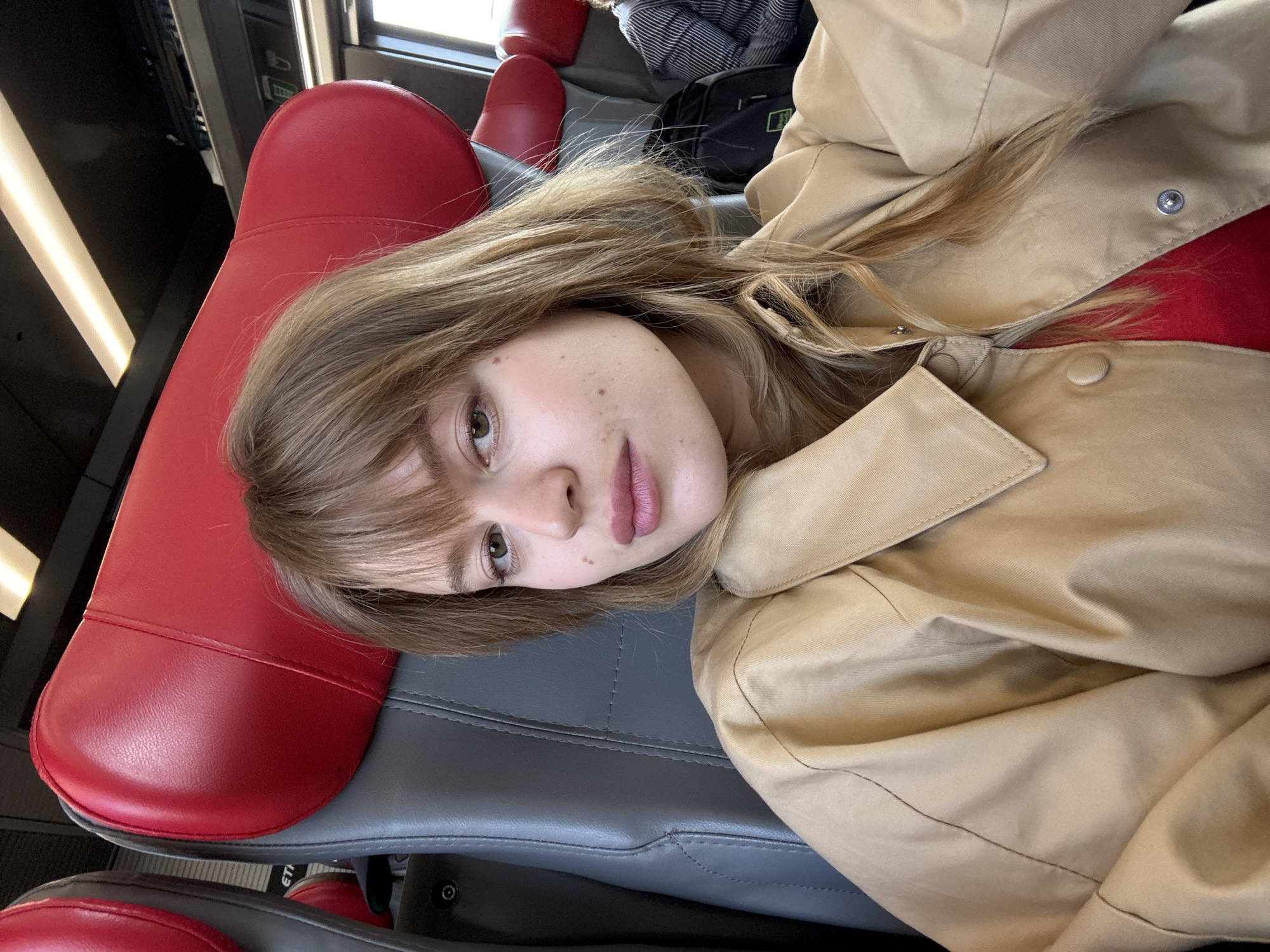 A close-up portrait shows a young woman resting inside a train, likely traveling in or around Mirano, Italy. She is positioned with her head gently angled, looking directly towards the camera with a relaxed, soft gaze. Her light brown hair, styled with bangs, frames her face, and her eyes appear light in color, possibly hazel. A few natural moles or freckles are visible on her cheek. Her lips are slightly parted, showing a subtle pink shade.
She is wearing a light tan or beige jacket or trench coat, with a hint of a bright red garment visible beneath, adding a touch of color to her attire. The setting is clearly a passenger train, identifiable by the distinctive red and grey padded seats. One bright red seat portion, possibly a headrest, is prominent near her head. In the background, a narrow, rectangular light fixture provides interior illumination, and a dark bag is partially visible on an adjacent seat. The ambient lighting suggests it could be daytime, though the exact time is not definitive. No specific visible text is discernible on the seats or in the background. The scene conveys a sense of calm and leisure during travel.