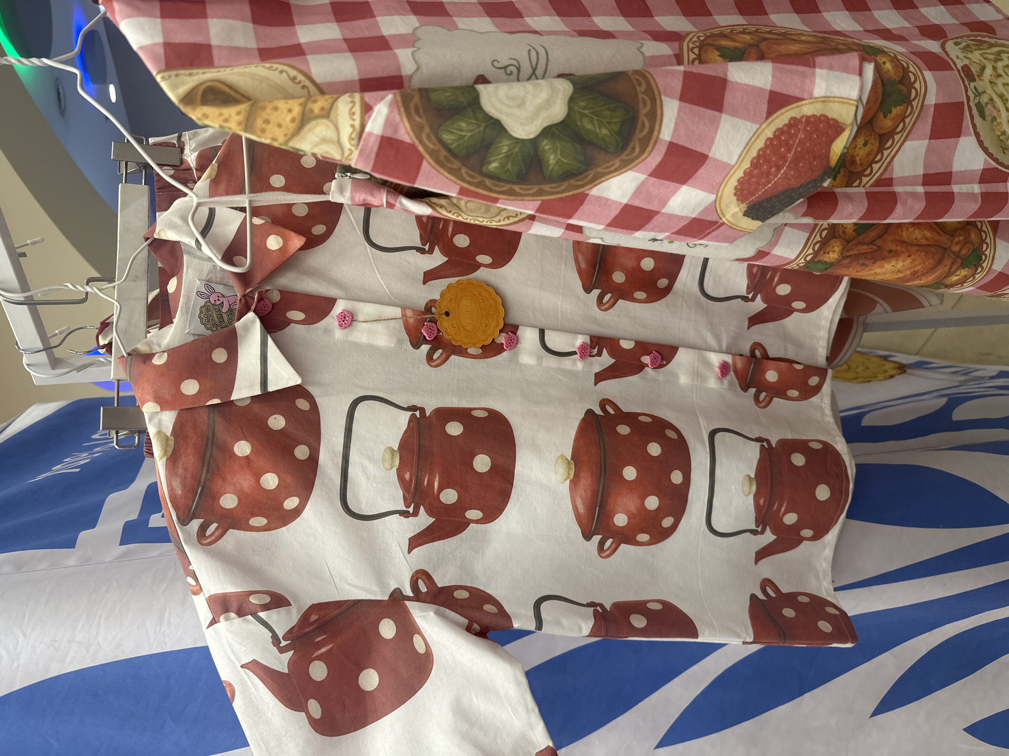The image shows a stack of hanging fabric items. The top item is a red and white gingham patterned fabric with images of prepared food items on plates. Beneath that is a white fabric with a repeating pattern of red polka-dotted teapots. The fabric is gathered at the top with a string of small pink buttons. A yellow circular tag is attached. The fabrics are hanging from a metal drying rack. The background appears to be a bed with a blue and white patterned sheet. The location is unknown.