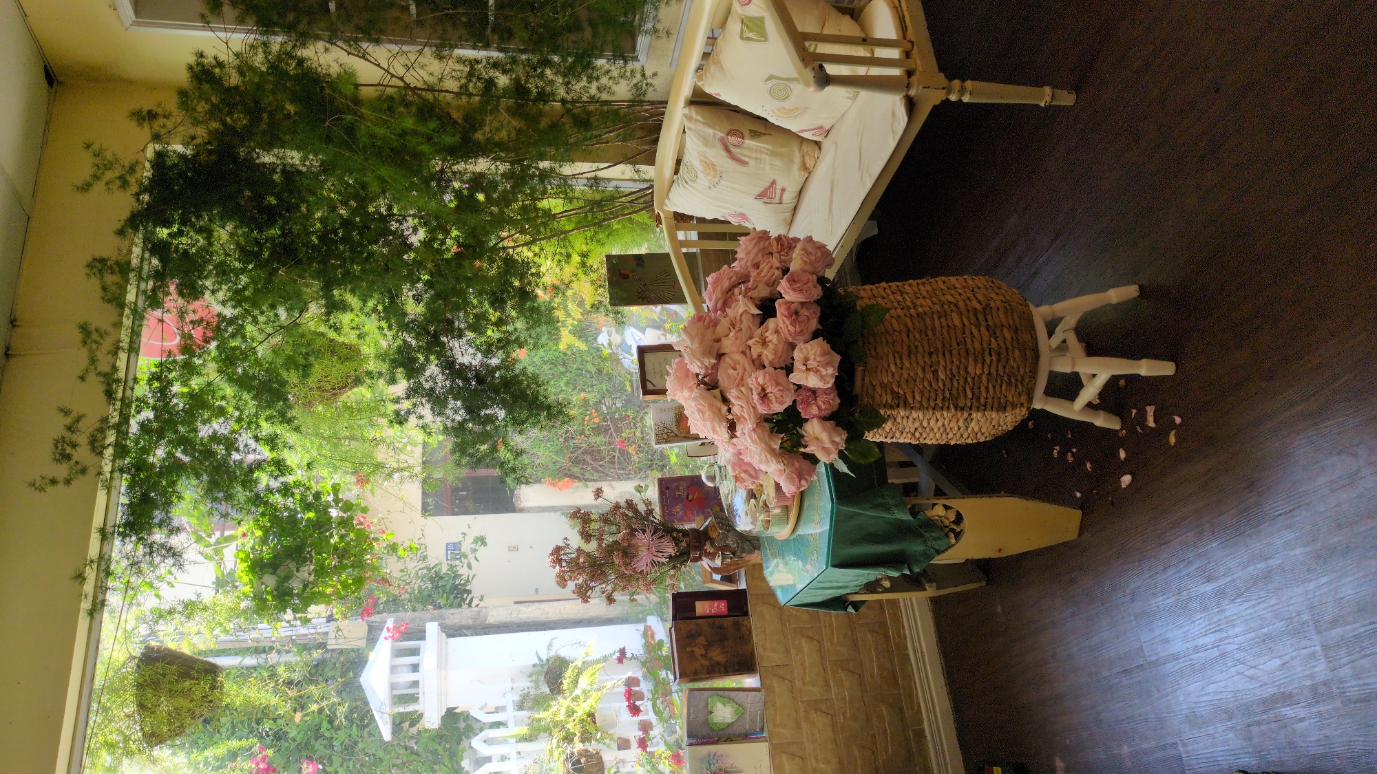 The image depicts an interior scene in Da Lat, Vietnam. A wicker basket filled with pink roses sits on a white wooden stand. To the left, a window overlooks a garden with a white picket fence. A white bench with cushions is partially visible in the upper right corner. A small table with a green cloth and books is also present. The floor is dark wood. The lighting is bright, and the overall atmosphere is soft and floral.