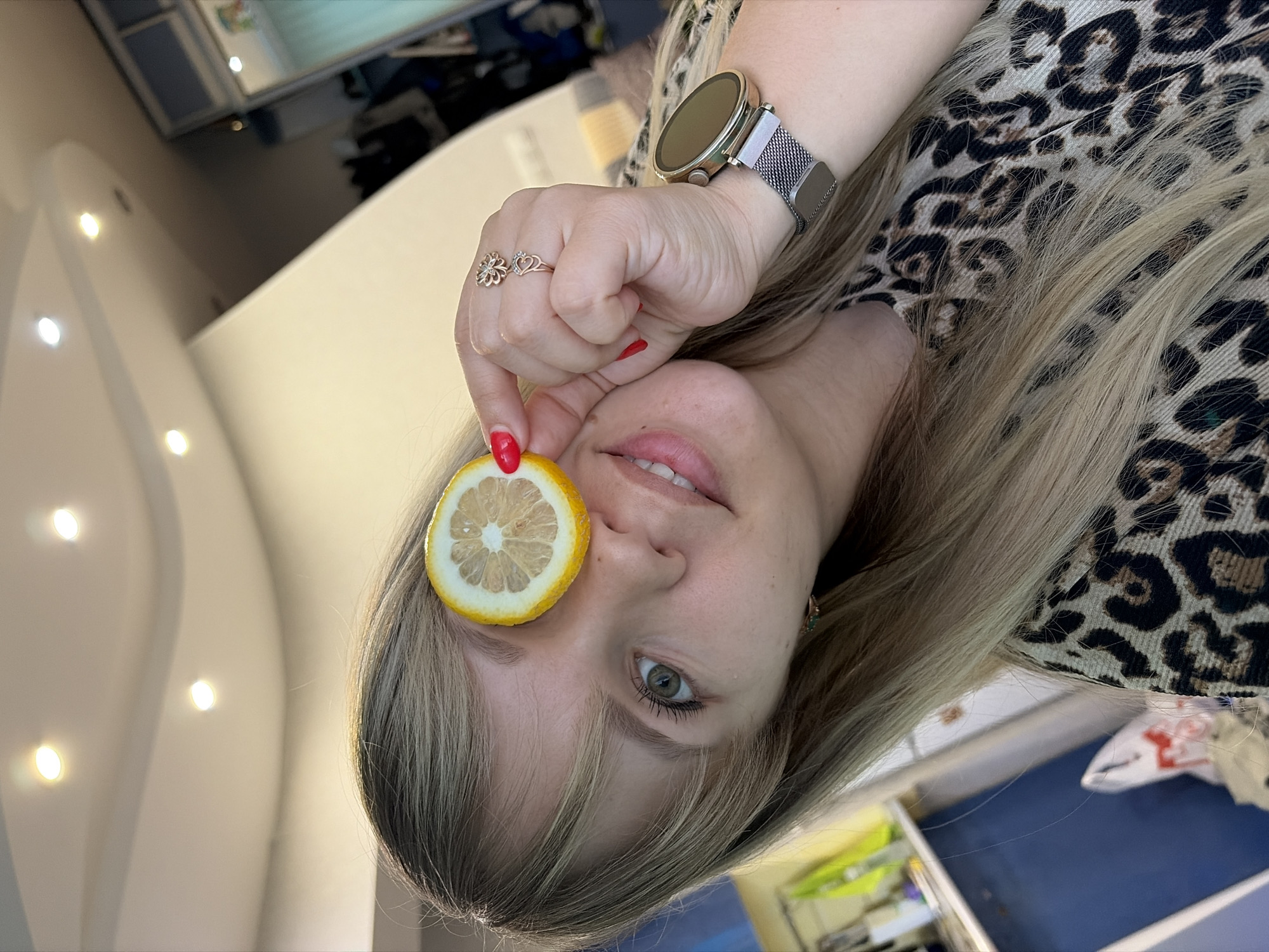 A woman with blonde hair and a leopard print shirt is holding a slice of lemon in front of her eye. She is wearing a gold watch with a silver band on her wrist, and a flower-shaped ring on her finger. The woman's fingernails are painted red. The location appears to be an interior space, possibly a residence or office, but the details are not clear enough to confirm the city or any specific location within Chaikovsky, Russia.