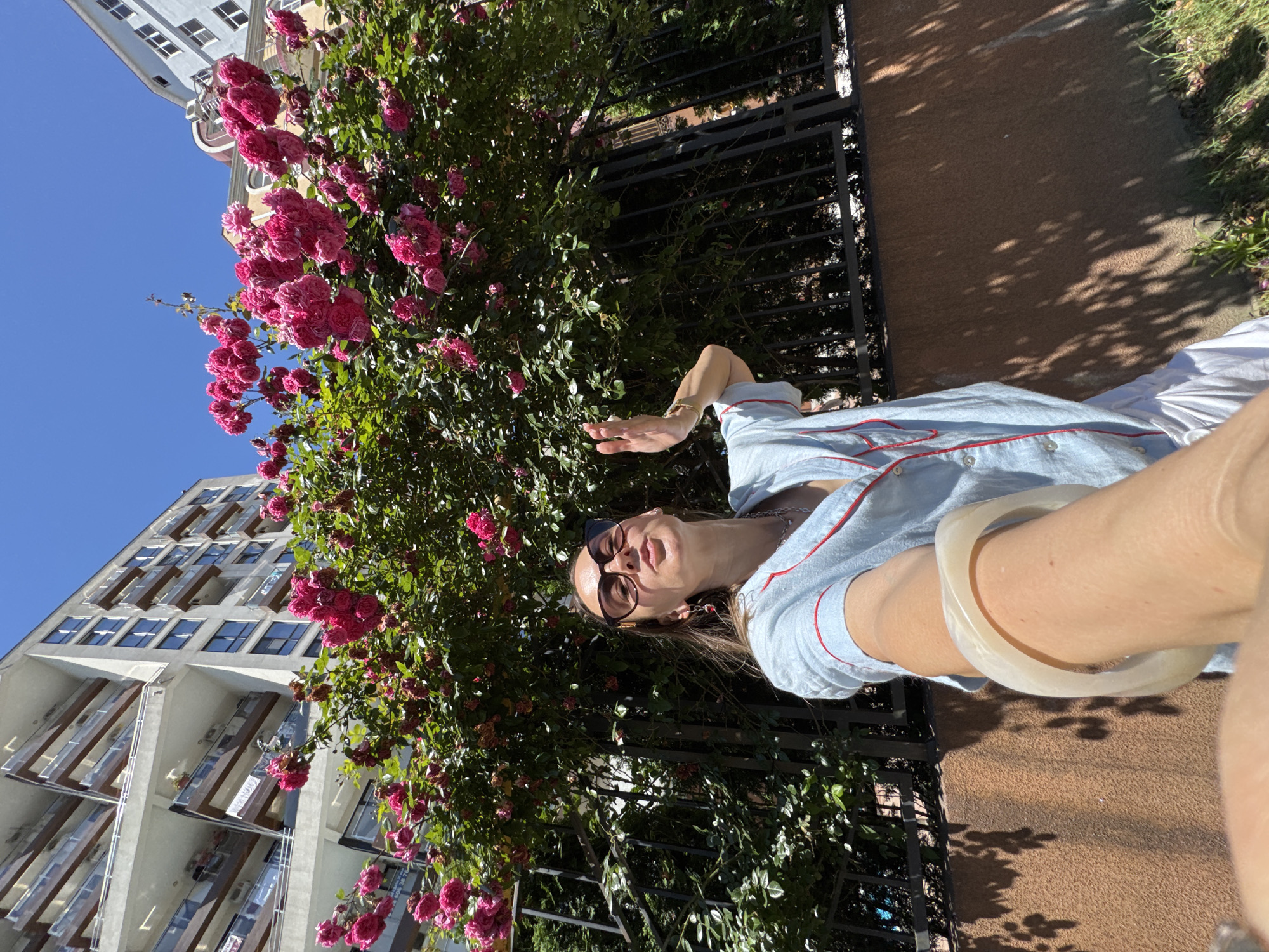 A young woman is taking a selfie outdoors in a city. She is standing in front of a flowering rose bush and a modern apartment building. The woman is wearing sunglasses, a light blue pajama-style top with red trim, and white shorts. She has a large white bracelet on her left wrist and is wearing earrings. Her right hand is raised, and she is puckering her lips. The apartment building is white with balconies and dark-framed windows. The rose bush is full of vibrant pink flowers and green leaves. The scene appears to be in Sochi, Russia, given the architectural style of the building and the lush vegetation. The sky is clear and blue. The ground is covered with grass and a brown wall with dappled shadows from the sun.