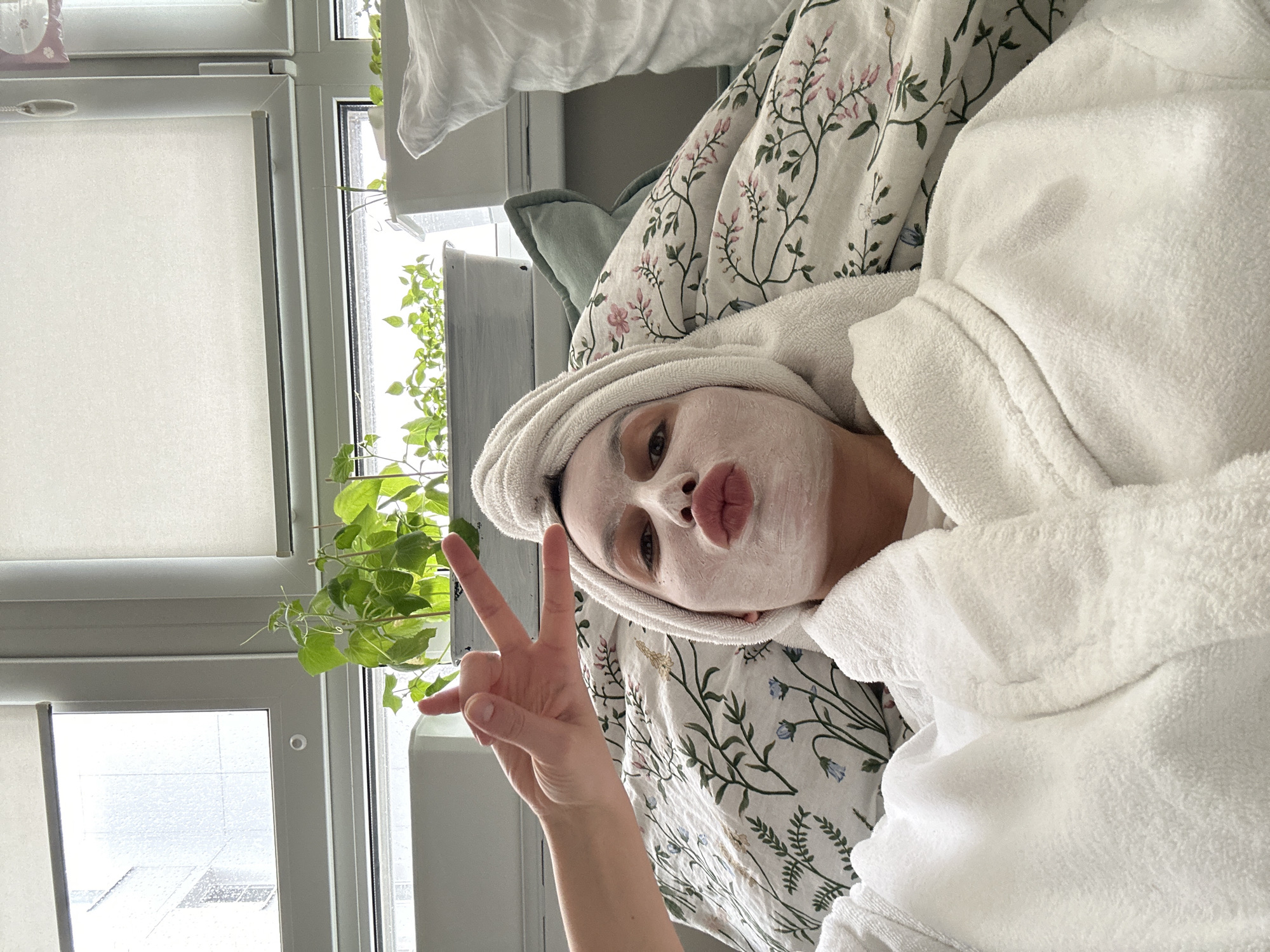 This tilted selfie captures a young adult, likely female, relaxing indoors, possibly in a bedroom in Moscow, Russia. She is lying down, facing the camera, with a white face mask applied to her face and a white towel wrapped around her head, suggesting a self-care activity. She wears a white, fluffy bathrobe. With her left hand, she makes a "peace" sign towards the camera, while her lips are playfully pursed in a "kiss face" expression. Her eyes are visible above the mask, looking directly at the viewer.
She is resting on a bed adorned with floral-patterned bedding featuring pink and green motifs, alongside a light green pillow. In the background, a large window with white roller blinds is visible, partially open. Green, vine-like plants are growing near the window, adding a natural element to the scene. Through the window, an urban environment with rooftops under an overcast or cloudy sky can be seen, indicating it is daytime. The overall mood is relaxed and playful. There is no visible text in the image.