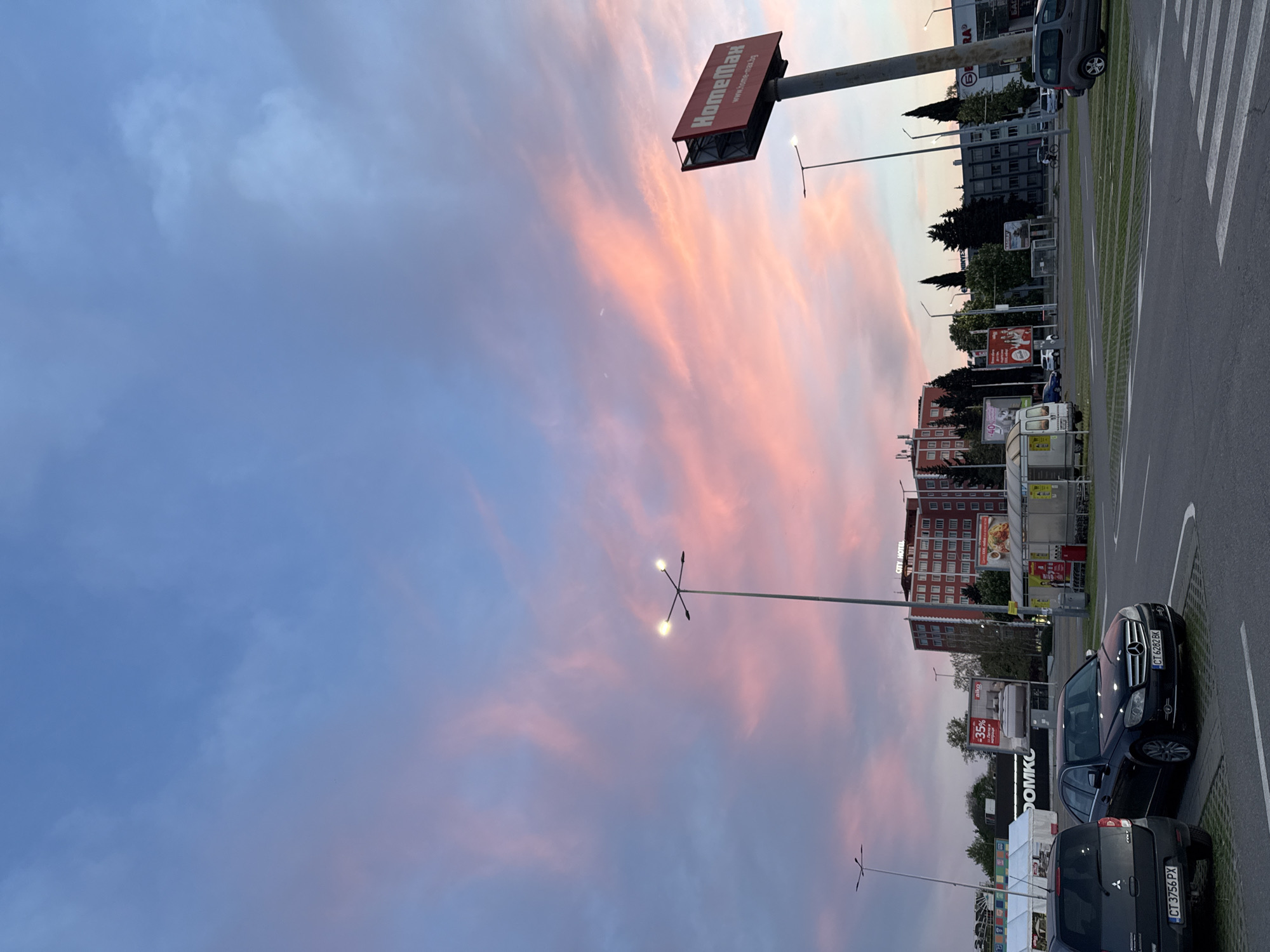 Here is a description of the image:
The image captures an outdoor scene, likely a parking lot adjacent to commercial establishments, under a sky painted with the hues of twilight or sunset. The sky displays a gradient of colors, transitioning from a muted blue to pink and orange shades. A slender crescent moon is subtly visible.
On the right side of the image, cars are parked in designated spots. A black vehicle has license plate "CT 3756 PX". Another black car in front of it with license plate "CT 6282BK". The lot seems to be marked with painted lines, indicating parking spaces.
In the background, commercial buildings are visible. A tall sign with the word "Нометан" in red letters stands prominently. A sign below says, "www.home-max.bg". Another building has the words "CITY HOTEL" written on it. There is also a shop called DOMKO, where the name "aiko" can be read.
The presence of street lights, some with their lamps illuminated, suggests the approach of night.
Based on the presence of the HomeMax sign, the location is Stara Zagora, Bulgaria. The image is taken outdoors, in what seems to be a parking lot shared by different commercial establishments. The lighting indicates that it is either dusk or dawn.