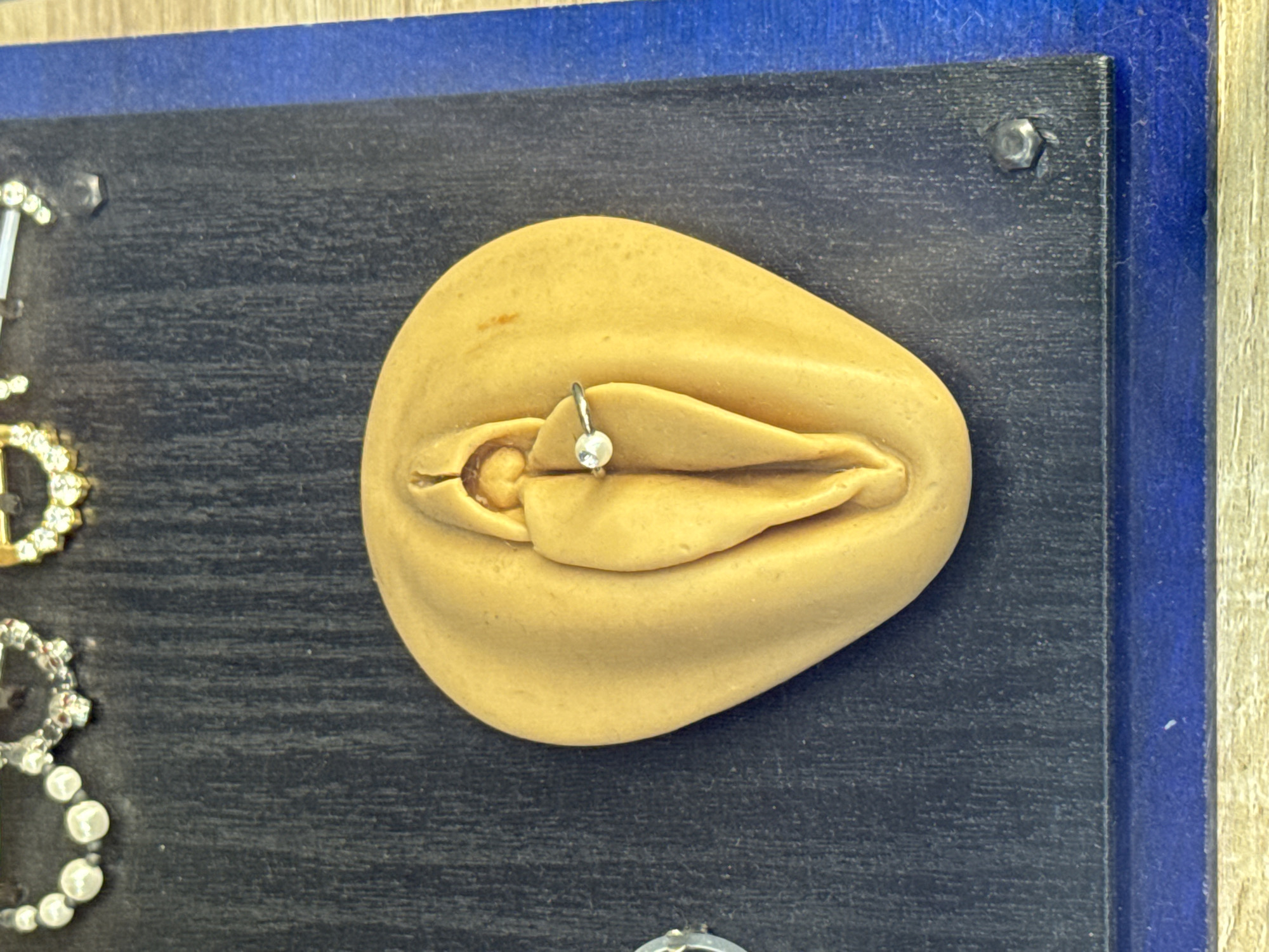 This is a close-up, eye-level, vertical shot of a sculpted object resembling a vulva. The object is light tan in color and appears to be made of a soft material like clay or dough. It is detailed with labia folds and a clitoral hood. A small, silver-colored ring with a pearl-like bead is pierced through the labia. The sculpted object is placed on a dark gray textured surface, which is bordered by a blue surface. In the background, partially visible at the top of the frame, are several pieces of jewelry, including pearl-like beads and sparkling stones. The image does not provide any information about the city of Lucerne, Switzerland, or any specific location.