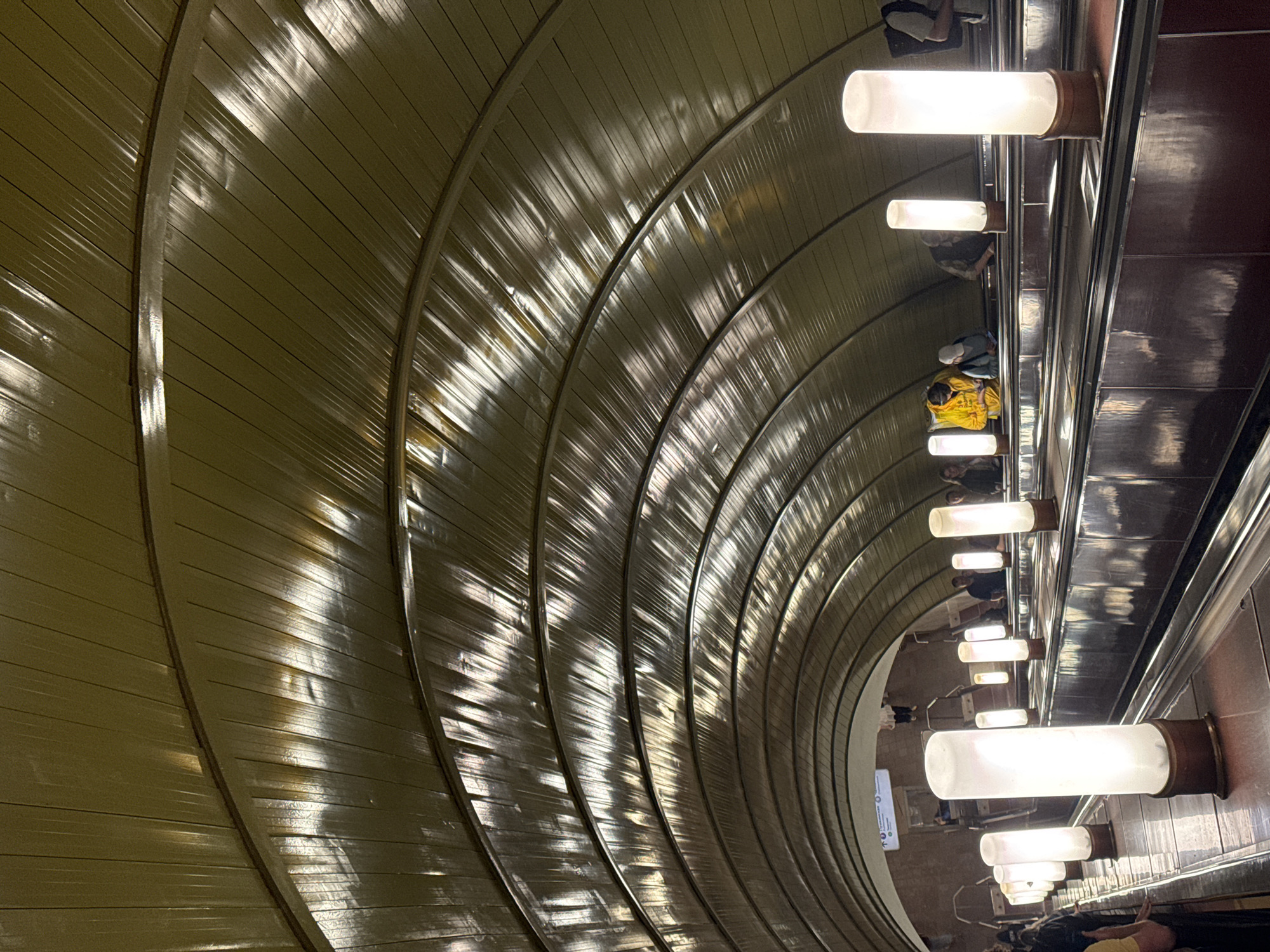 This media file depicts the interior of a deep metro station escalator tunnel, located in Moscow, Russia. The image is oriented sideways, giving the impression of looking across a steep, curving incline.
The most striking feature is the arched ceiling and wall on the left, composed of numerous ribbed, olive-green or dark metallic panels. These panels exhibit strong reflections from the artificial light, creating dynamic patterns of light and shadow that highlight the tunnel's impressive curvature and depth.
Along the right side of the escalator, a series of cylindrical, frosted white light fixtures are mounted horizontally. These lights are attached to a dark, reflective metal panel that runs parallel to the escalator steps, illuminating the path.
Several individuals are visible on the escalator steps. Their figures are partially obscured, but some appear to be standing, while one person wearing a bright yellow jacket is distinctly seen sitting on a step. Their presence indicates ongoing travel within a public transportation system.
At the far end of the long tunnel, a small, white rectangular sign with indecipherable text is faintly visible. The scene captures the unique architectural style and functionality of a deep underground metro environment.