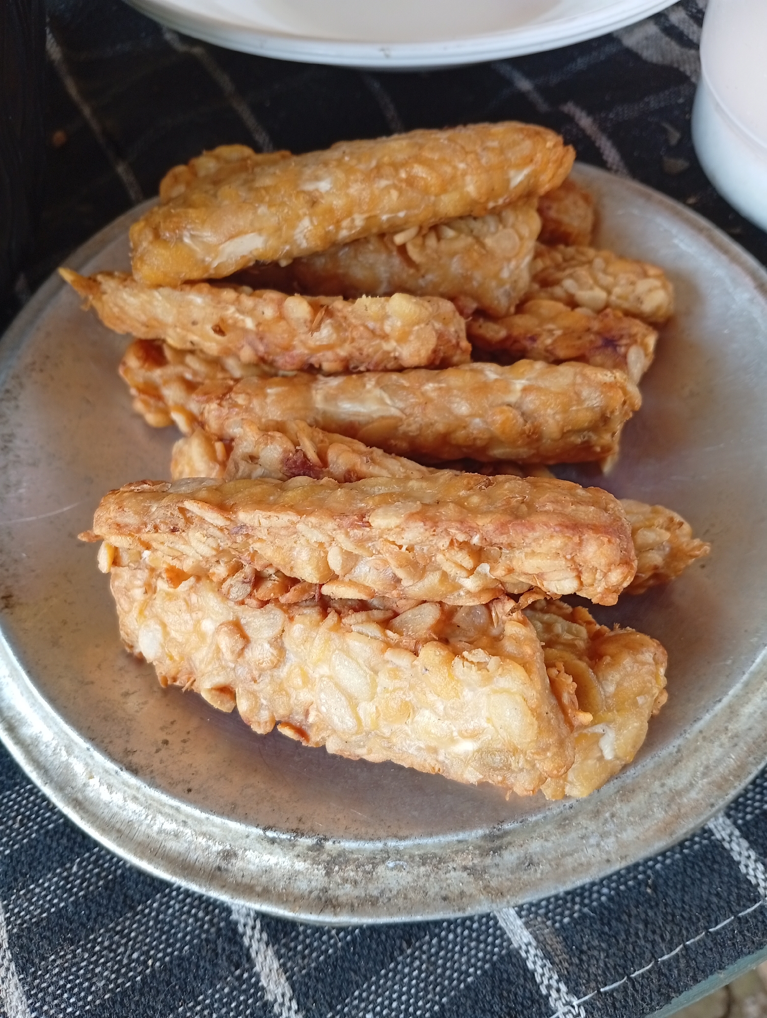 A close-up, overhead shot shows a pile of golden-brown, fried food items, possibly tempeh or banana fritters, arranged on a metallic, circular plate. The food items are oblong in shape and appear to be coated in a crispy batter. The plate sits on a dark fabric tablecloth with a checkered pattern of black, white, and grey lines. The background is blurred, showing hints of white plates and possibly a white bowl. The image appears to be taken indoors, with natural light illuminating the food. The scene is likely set in a home or a local eatery in Cisitu, Indonesia, given the context of the prompt.