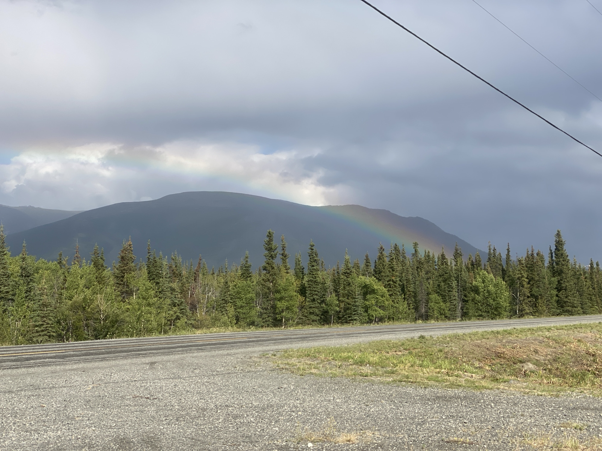 The image shows a highway in Haines Junction, Canada, with a dense forest of evergreen and deciduous trees lining the side of the road. In the background, a mountain range is visible under a cloudy sky, with a rainbow arching across it. The road is paved and appears to be a two-lane highway with a gravel shoulder. The sky is overcast, with patches of lighter clouds and darker storm clouds. The overall scene suggests a natural, somewhat wild environment, possibly after a rain shower, given the presence of the rainbow. There are no people or vehicles visible in the image.