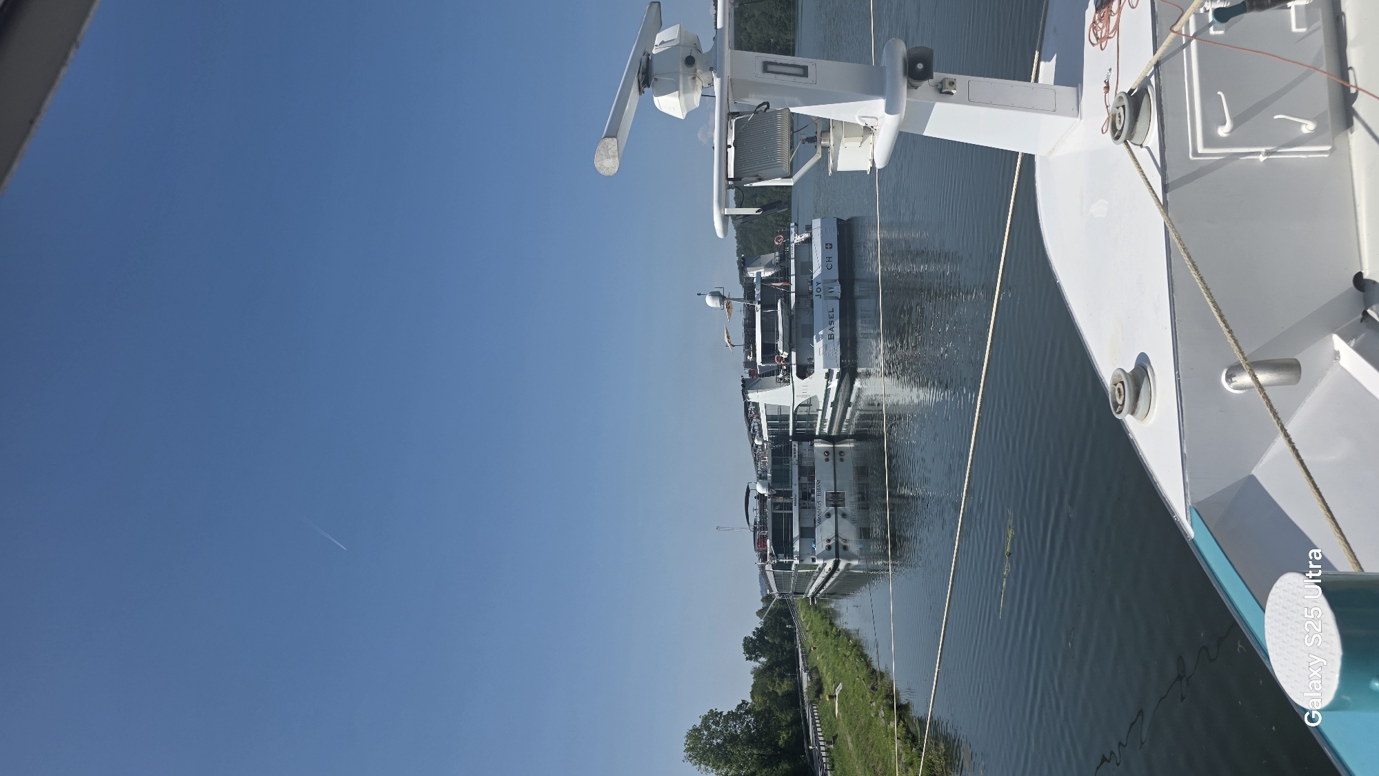 The image shows a clear blue sky with a faint contrail from an airplane. Below the sky are two large white river cruise ships docked side-by-side on a calm river. The ship on the right has "JOY BASEL CH" printed on its hull, and the ship on the left has "AMADEUS THENE" printed on its hull. The ships are moored in what appears to be a river port in Passau, Germany, as indicated by the presence of the Swiss flag on the "JOY" ship and its name referencing Basel. The riverbanks are lined with green grass and trees. In the foreground, the bow of another white boat is visible, equipped with nautical equipment like a radar antenna and cleats. The water is reflective, mirroring the sky and the ships.