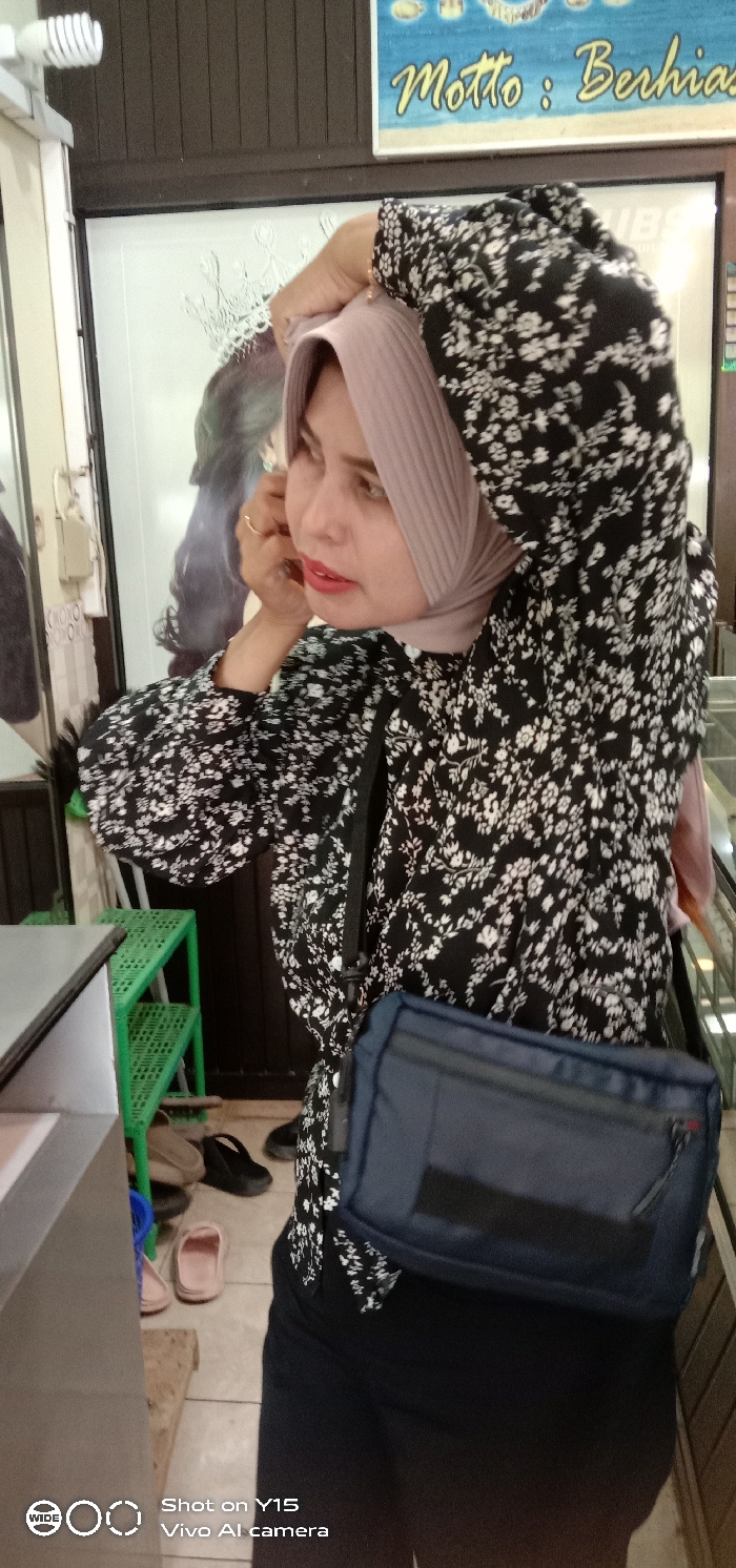 The image depicts an indoor commercial setting, specifically a shop in Maron, Indonesia. A woman is positioned prominently, facing towards the viewer's right, with her torso slightly angled. She is wearing a light purple ribbed hijab, a black long-sleeved shirt with a white floral pattern, black trousers or skirt, and a dark blue messenger bag slung across her body. Red lipstick is visible. Her right hand, adorned with a gold-colored ring on the ring finger, is raised near her ear, suggesting she is adjusting her hijab or an earring. Her gaze is directed towards the upper-right.
In the background, a light-colored wall features a mounted electrical box and wiring. To the woman's left, a reflection of a crown design is visible on a glass surface, potentially a display case. Below this, a green three-tiered plastic rack is present, and several pairs of shoes, including brown loafers, black flats, and pink slippers, are scattered on the tiled floor. In the upper-right background, a sign with a blue background and yellow text is partially visible, reading "Motto : Berhias" (Motto: Decorate/Beautify), indicating a beauty or jewelry-related business. Faint reflections of the letters "UBS" are also discernible on a nearby glass surface. A white LED light bulb fixture is mounted in the upper-left corner. A light-colored counter or display case is partially visible in the lower-left foreground.