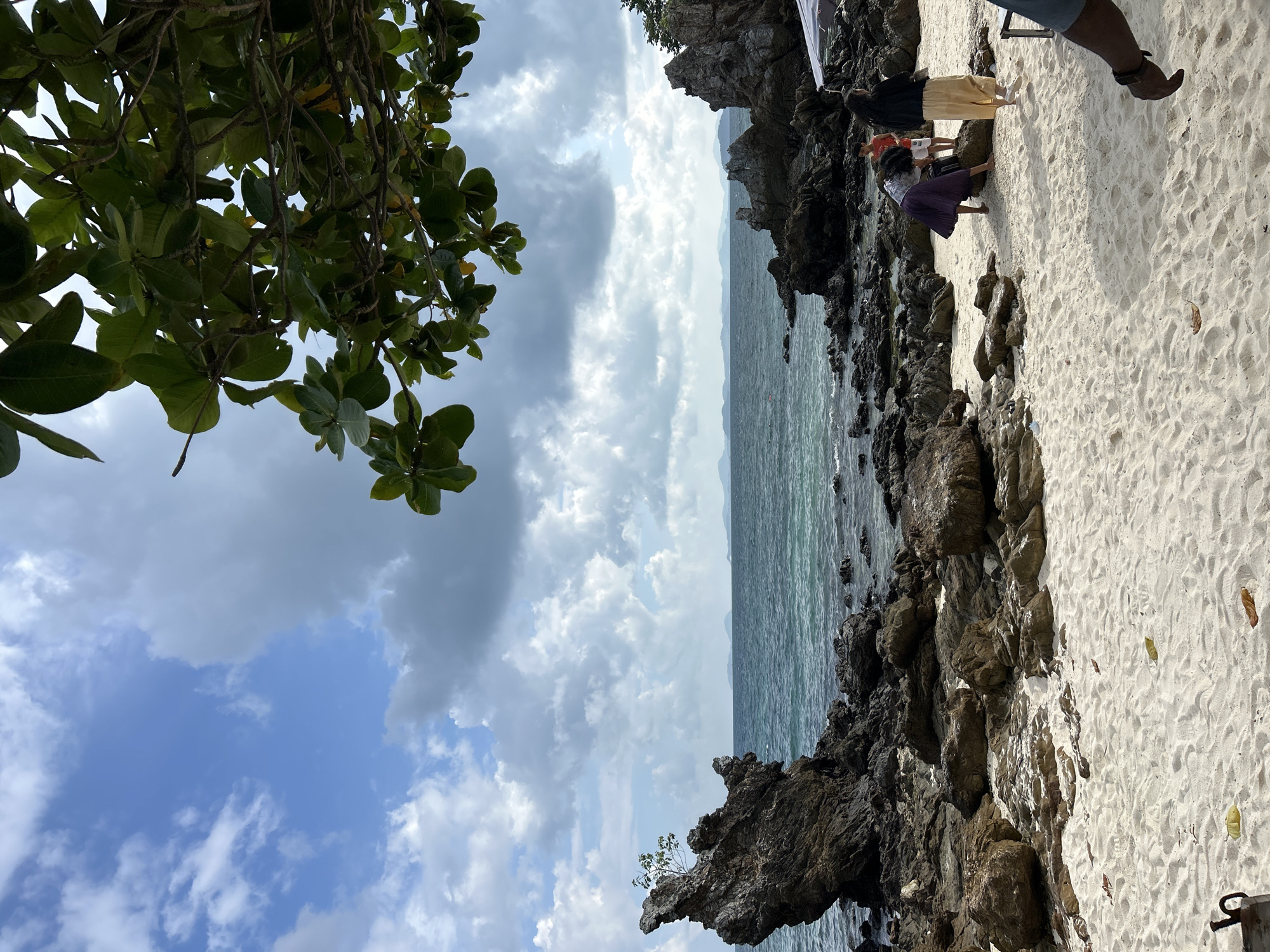 A scenic view of a rocky beach in Phru Nai, Thailand, is captured under a partly cloudy sky. The foreground features a sandy beach with scattered dry leaves and a metallic object in the lower left corner. In the mid-ground, several people are present. Two individuals, appearing to be women, are interacting with the rocks near the water's edge. One is wearing a purple skirt and bending down, possibly picking something up, while the other, in a beige skirt and dark top, stands nearby. A younger person, a boy in an orange shirt and white shorts, is also visible. The middle ground is dominated by dark, jagged rocks that extend into the turquoise ocean. A distinctive tall, sharp rock formation stands prominently on the left side of the image, with a small tree growing precariously from its top. The background shows the calm sea stretching towards a hazy horizon with distant islands. Lush green foliage from a tree hangs into the upper right corner of the frame, providing a natural canopy.