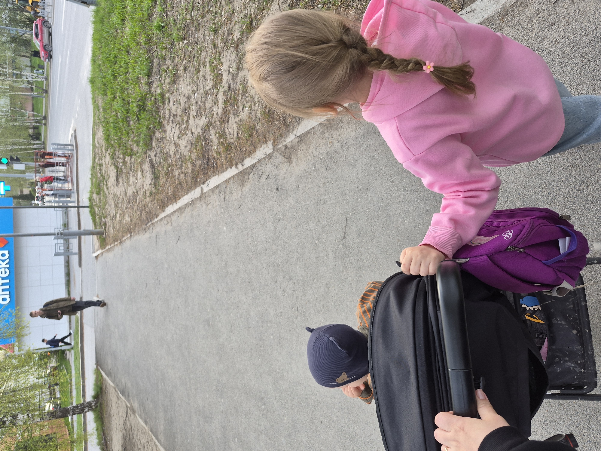 A medium-angle shot, taken from above, shows a girl in a pink hoodie next to a stroller being pushed by someone wearing black, likely on a sidewalk in Tobolsk, Russia. The girl is facing the stroller, possibly looking inside. The stroller holds a purple backpack. In the background, across the sidewalk, are buildings and trees, with a blue and white sign visible, reading "аптека" ("pharmacy" in Russian). Two men are walking away from the viewer in the distance, with a red car visible further down the street. The setting appears to be an urban area, captured during daylight.