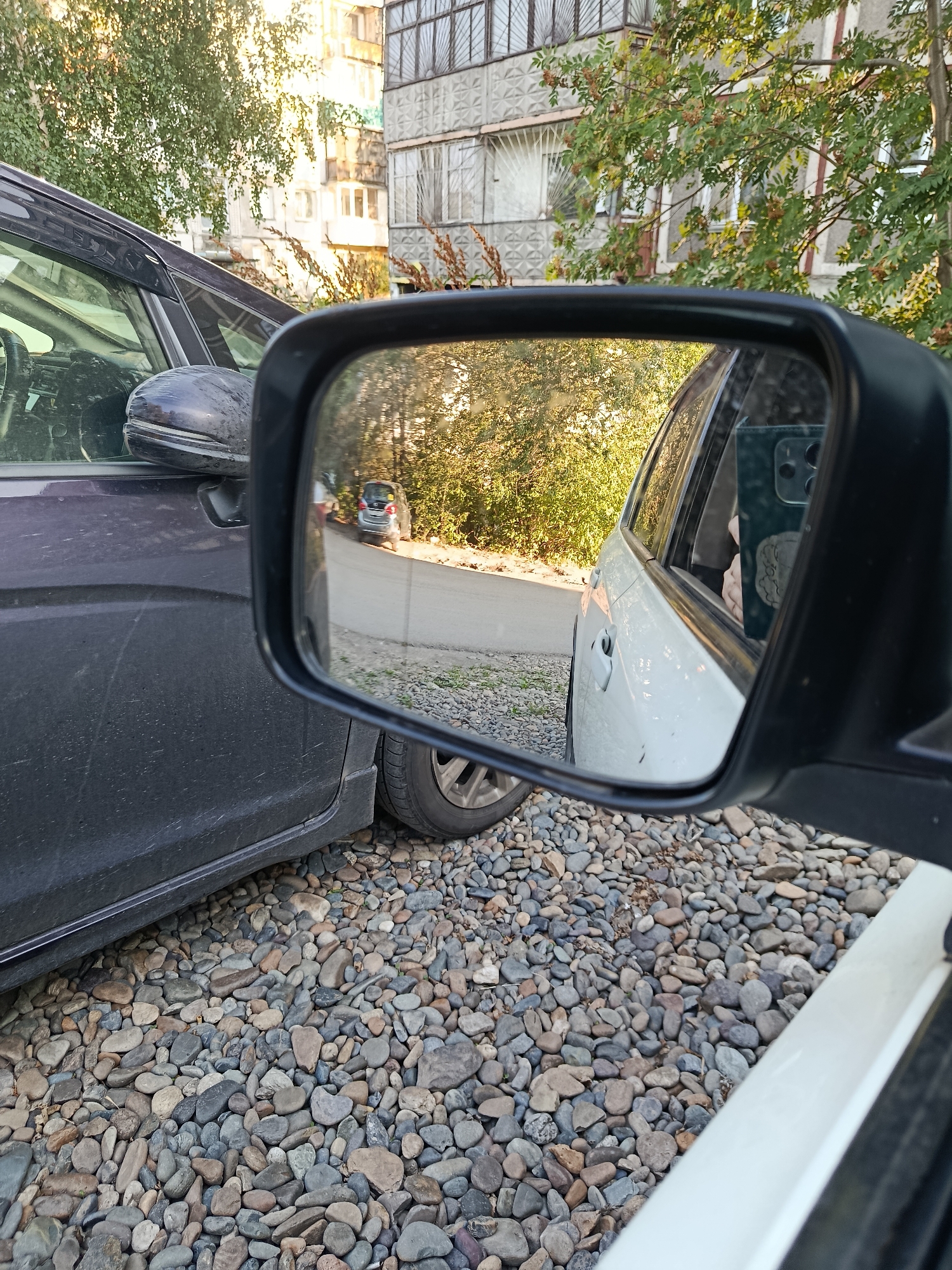 The image shows a side-view mirror reflecting a street scene in Biysk, Russia. The mirror reflects a light-colored car parked on the right, with a person holding a phone inside. The street is paved with asphalt and lined with gravel on the left. A dark-colored car is parked on the left side of the image, partially obscuring the mirror. In the background, there is a multi-story building with concrete walls and a tree with green leaves and red berries. The sun is shining, casting shadows and highlighting the scene.