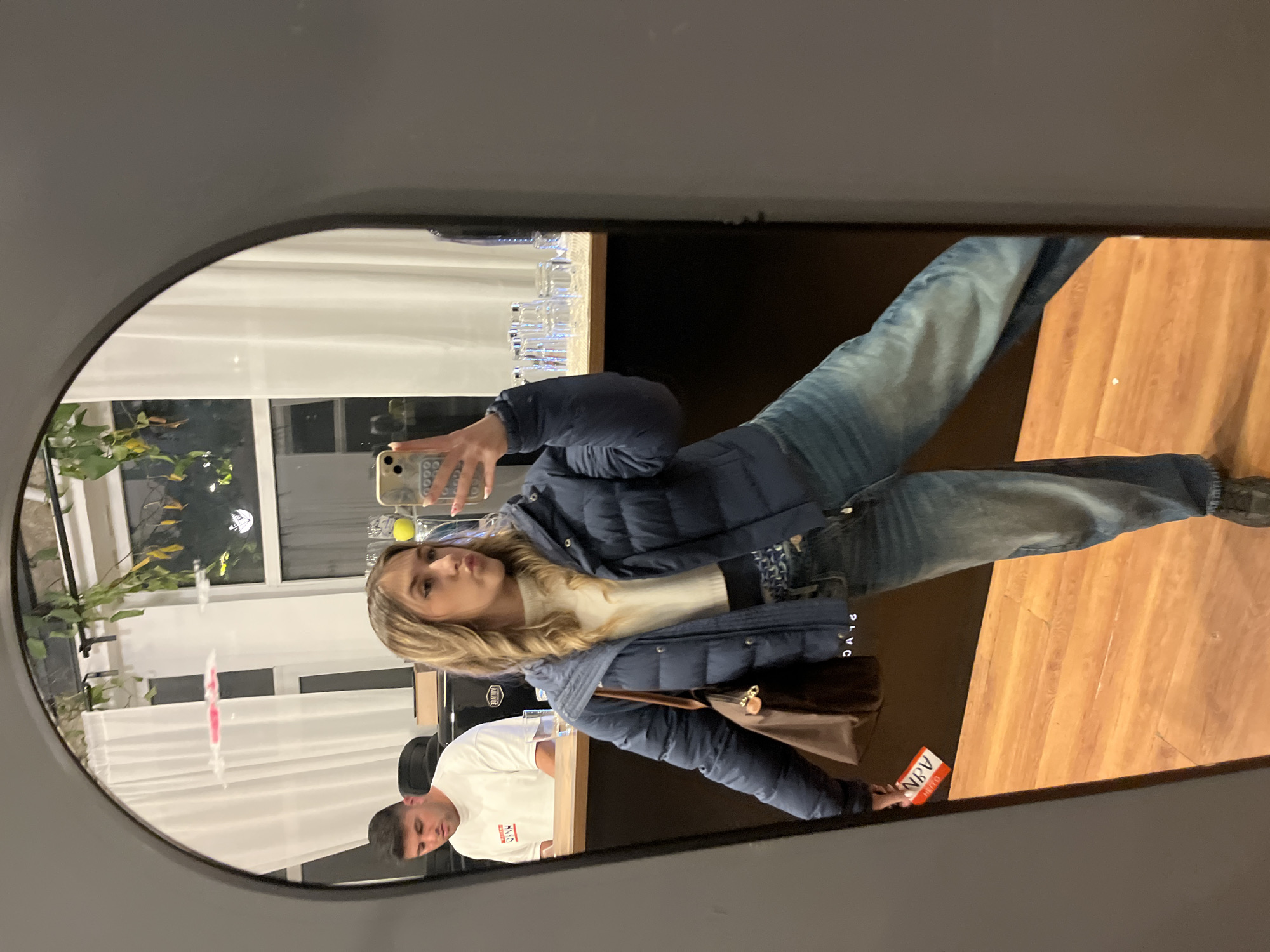 A young woman is taking a selfie in a mirror. She is wearing a navy blue puffer jacket, a white turtleneck sweater, and blue jeans. She has blonde hair and is holding a cell phone in her right hand. The mirror is oval-shaped and has a black frame. Behind her, a man is standing at a counter wearing a white t-shirt. There are glasses lined up on the counter behind him. A brown purse hangs from her left shoulder. The background shows a window with a white blind and some plants outside. A brown wooden floor is visible. The photograph was likely taken in Sofia, Bulgaria, as indicated by the "Hello ABV" sticker near the bottom right corner of the mirror, a local delivery company.