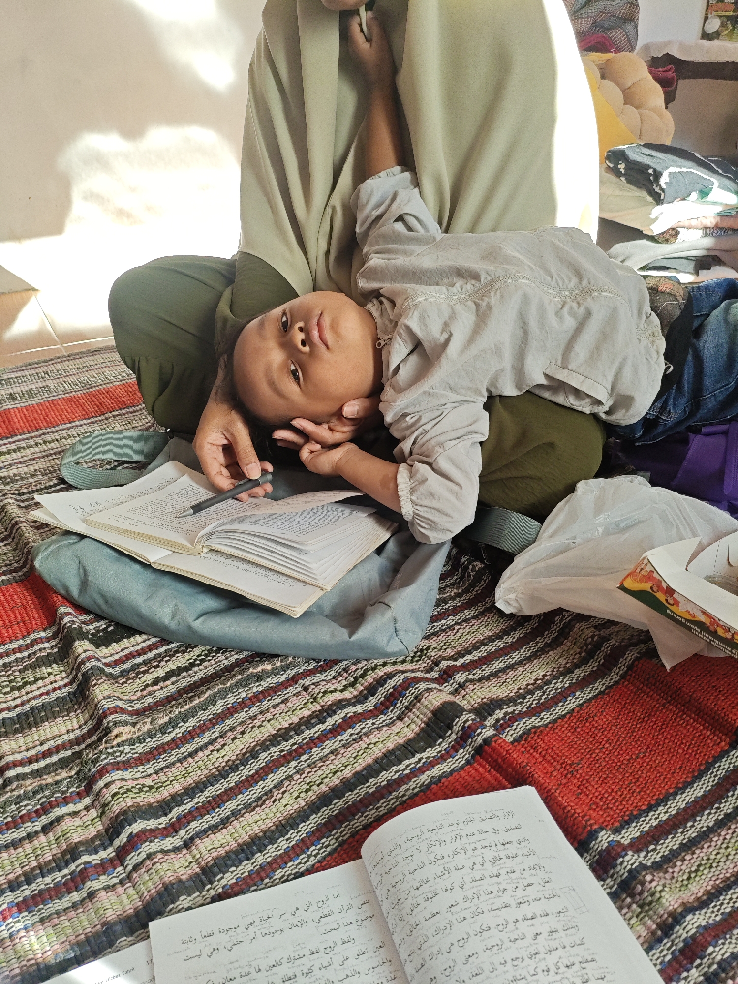 In Rejoagung, Indonesia, a young child with dark hair and eyes lies on their back, resting their head on their hand. They are wearing a light-colored shirt. The child is positioned on a patterned rug that features horizontal stripes in various colors, including red, black, white, green, and blue. In the foreground, an open book with Arabic text is visible, resting on a blue bag. A dark pen lies on the open pages of the book, held by a person whose legs are bent and covered by a green garment. Part of this person's arm is also visible, resting behind the child. In the background, a stack of folded clothing can be seen, including items of blue and purple. Sunlight casts shadows on a white wall in the upper left of the frame.