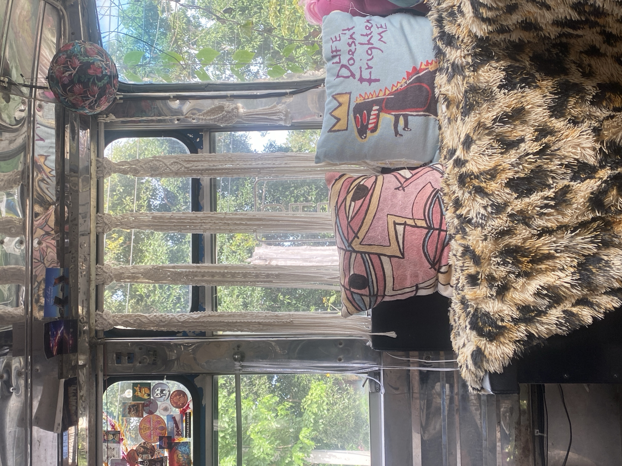 The image shows the interior of a vehicle, likely a bus or van, positioned in Moo 4, Thailand, judging by the view through the window. The interior is cluttered with personal belongings. A macrame window covering hangs over the window, partially obscuring the view of trees and foliage outside. A floral fabric ball is attached near the top of the window. Two cushions are stacked on top of each other: a light blue cushion with an illustration of a dinosaur and the phrase "LIFE Doesn't Frighten ME", and a pink cushion with geometric shapes. A faux fur blanket with a leopard print covers a portion of the cushions. The walls of the vehicle are metallic and covered in stickers.
