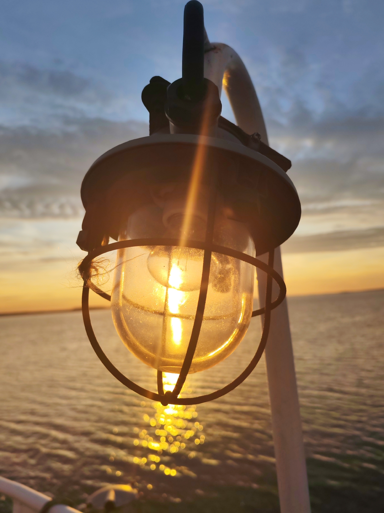 A close-up shot shows a nautical-style lantern casting a warm light against a sunset backdrop over a body of water. The lantern, enclosed in a protective metal cage, hangs from a white post. The water reflects the golden light of the setting sun, creating a glittering effect on the surface. The sky above the horizon displays a blend of blues and oranges.