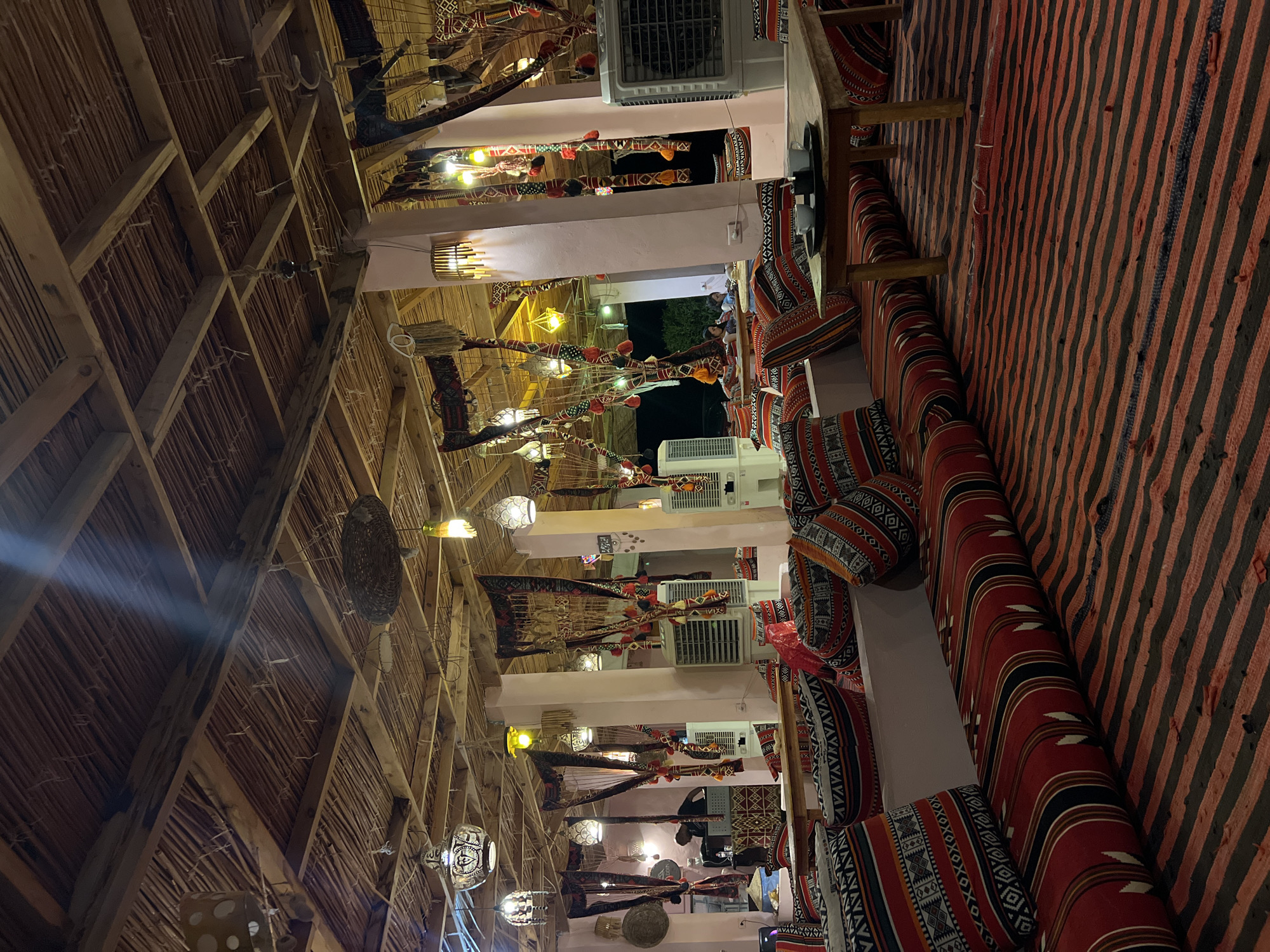 A low-angle, medium shot captures the interior of a Bedouin-style restaurant in Nuweiba, Egypt, at night. The ceiling, constructed of wooden beams and dried palm fronds, is adorned with numerous decorative lights and woven fabrics. Large, white evaporative coolers are visible along the walls, suggesting a warm climate. The seating area is furnished with floor cushions and banquettes upholstered in traditional striped fabrics of red, black, and white. A simple wooden table in the foreground holds two white cups on a black tray. In the background, a person is visible behind a counter, and a few patrons are seated at a table. The overall impression is one of rustic, open-air dining.