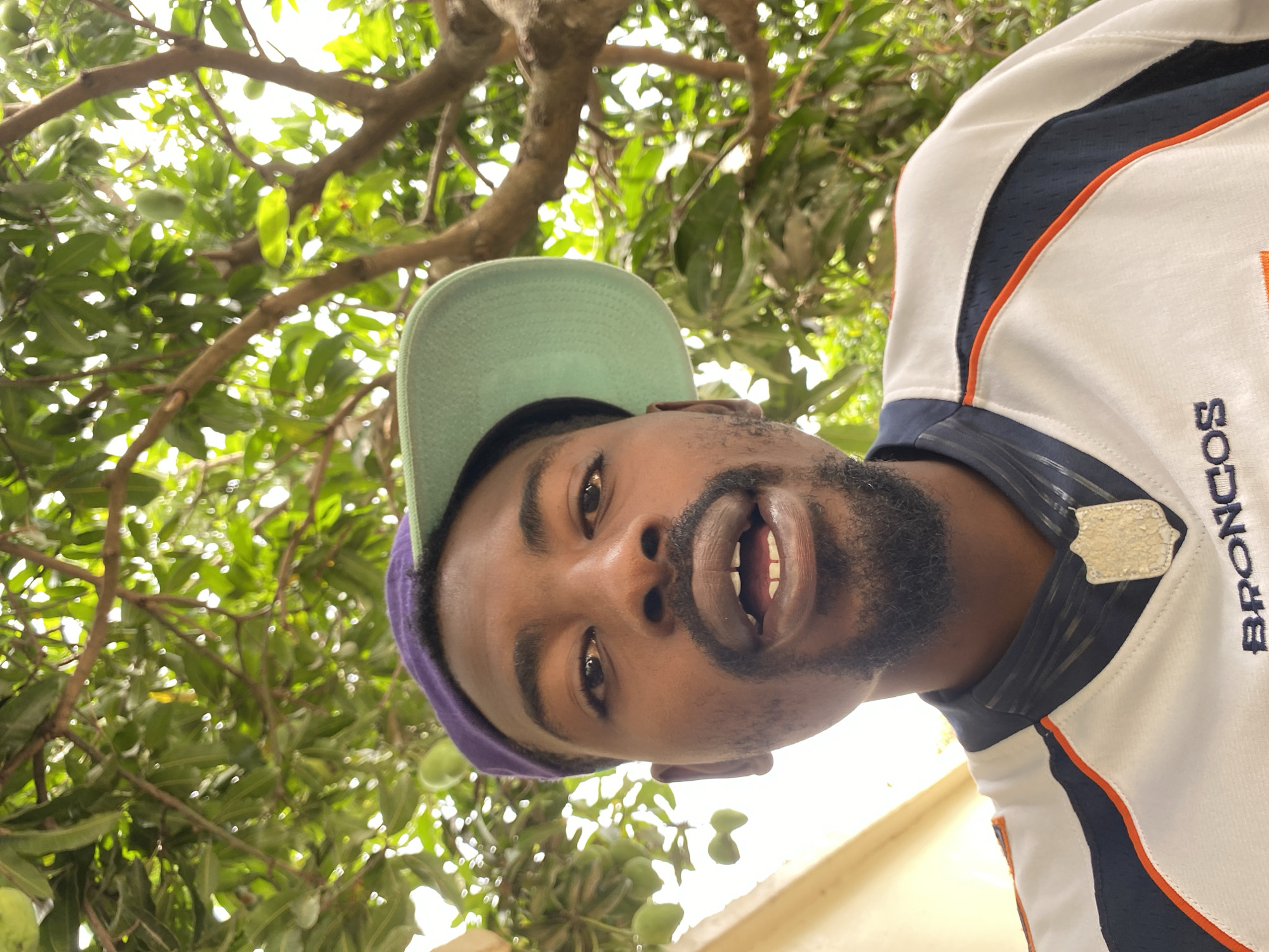 A close-up, tilted photograph shows a Black male wearing a light green cap with a purple underside and a Denver Broncos jersey. He is looking directly at the camera with his mouth open, revealing his teeth. He has dark eyes, a beard, and short hair. The background is filled with green foliage and tree branches. The location is Barakin Akawo, Nigeria.