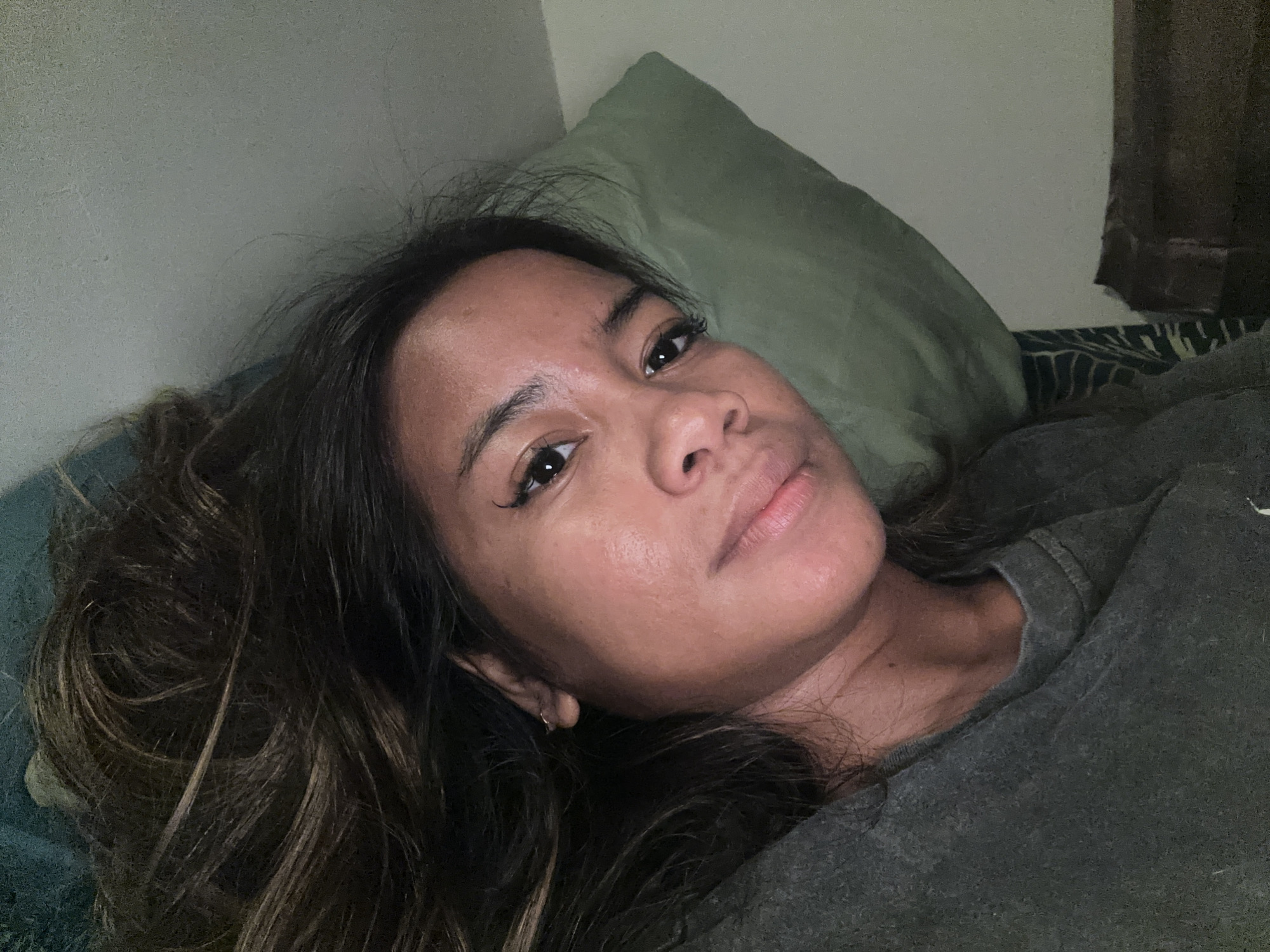 The image shows a woman lying on a bed, with her head propped up on a green pillow. She is looking towards the camera with a relaxed expression. Her hair is long and dark brown, falling around her face and shoulders. She is wearing a dark gray t-shirt. The background appears to be a bedroom, with a textured wall on the left and a dark curtain on the right. The bed is covered with a dark patterned sheet and a gray blanket. The lighting is soft and natural. There is no indication of the specific location within Sukawati, Indonesia, other than it appears to be an indoor residential setting.