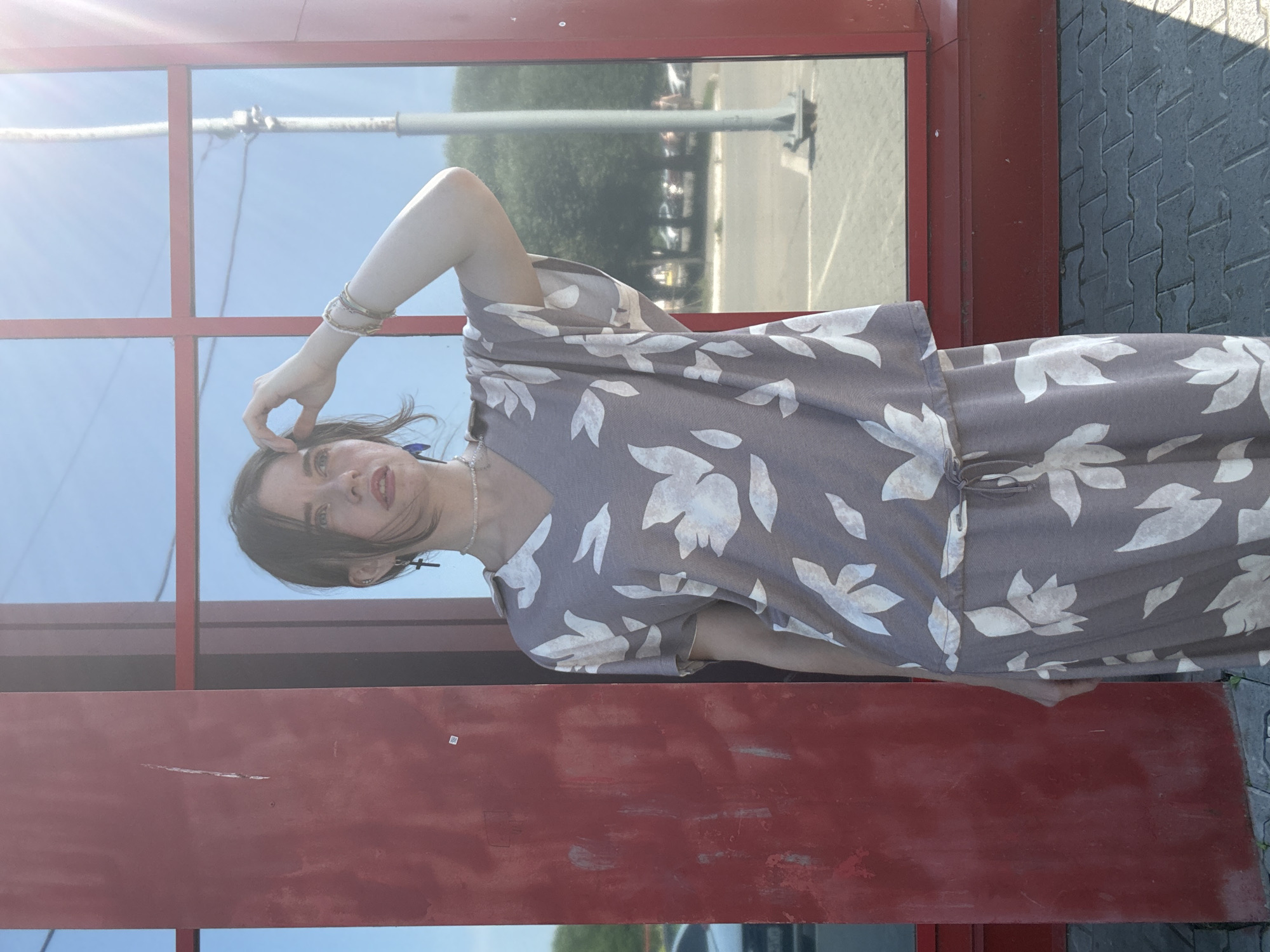 A young woman stands in front of a red-framed window on a sunny day. She is wearing a grey patterned dress with white floral motifs. The dress has a V-neck and short sleeves. Her hair is dark brown and pulled back. She is wearing a necklace and earrings. Her right arm is raised, with her hand touching her hair. The window reflects the blue sky and some trees. The ground is paved with grey bricks. The overall scene suggests an outdoor urban environment.