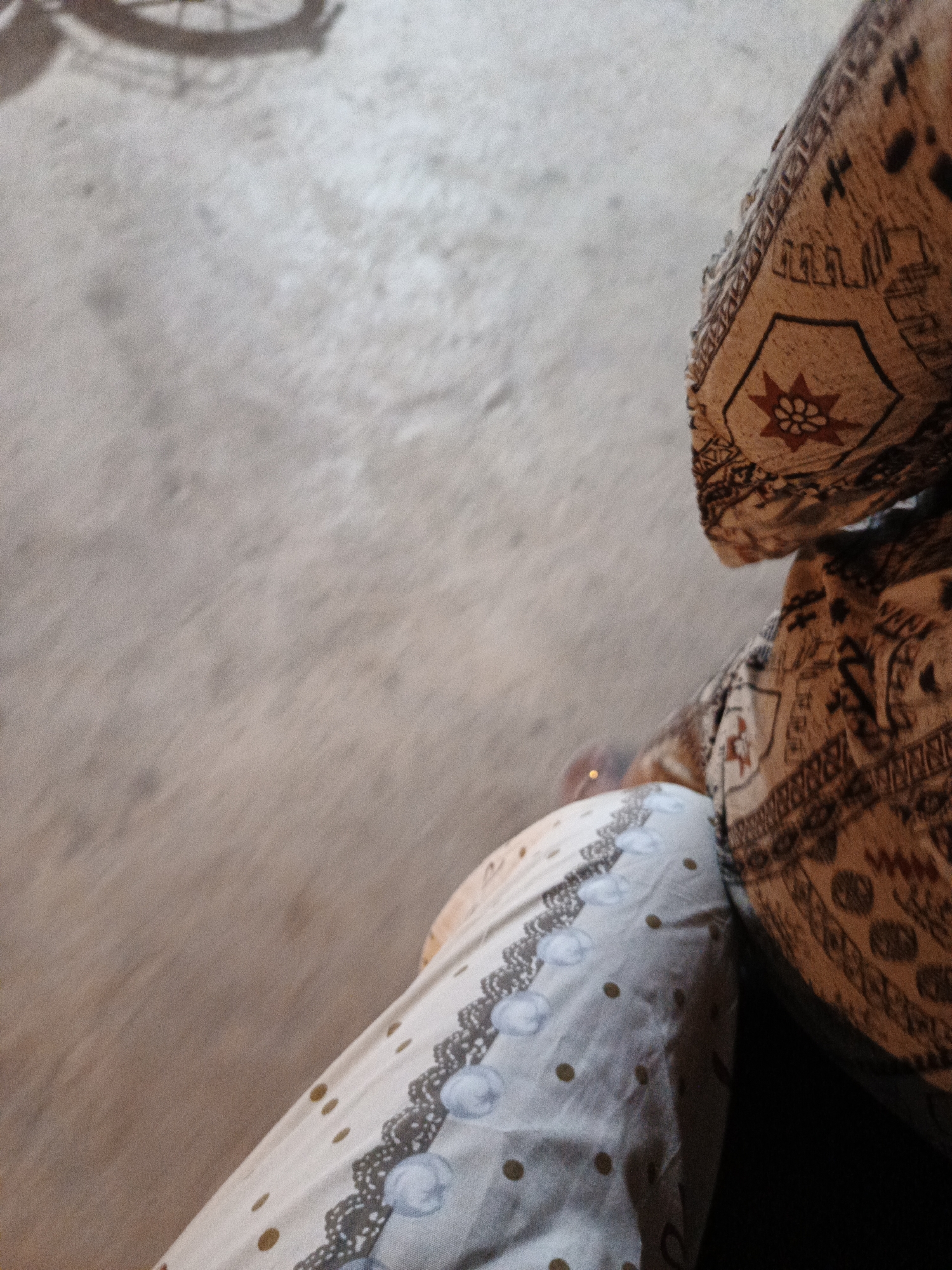 The image shows a close-up view of a person's legs, viewed from a high angle. The person is wearing patterned clothing. The background is a textured, light-colored surface, possibly concrete or asphalt, with a blurred shadow of a circular object, likely a bicycle wheel, visible in the upper left corner. The scene appears to be outdoors, on a street or pathway, in Sumbersari, Indonesia. There are no other people or animals visible in the frame.