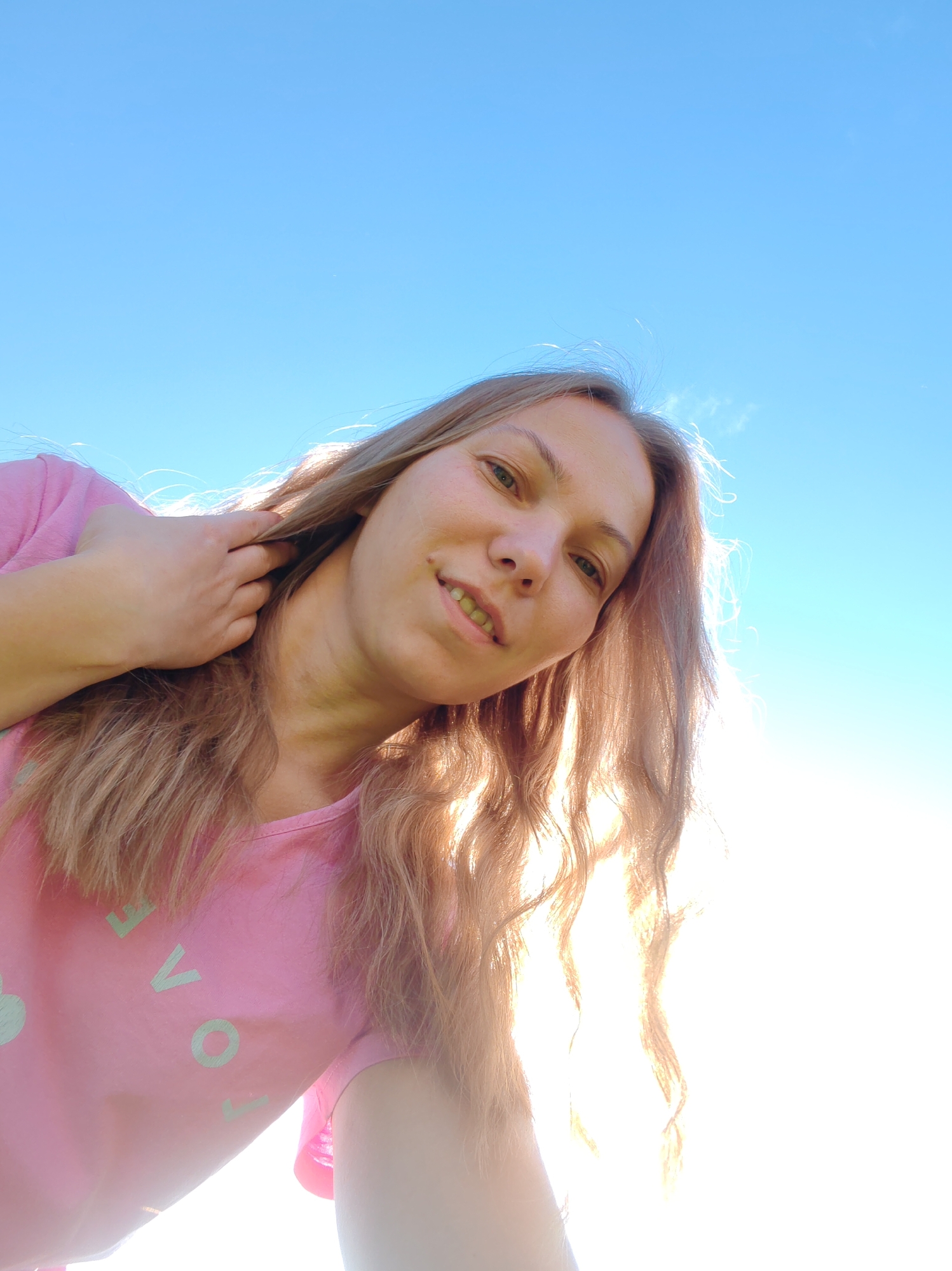 An upward-angle selfie shows a young woman with long, wavy blonde hair, wearing a pink t-shirt with the word "LOVE" printed in light green. Her hand is in her hair, and she is looking at the camera with a slight smile. The background is a clear, bright blue sky with a hint of sunlight. The image was taken outdoors, possibly in a park or open area, during the daytime. There are no discernible landmarks or architectural elements that would indicate a specific location within the city of Chukhlem, Russia.