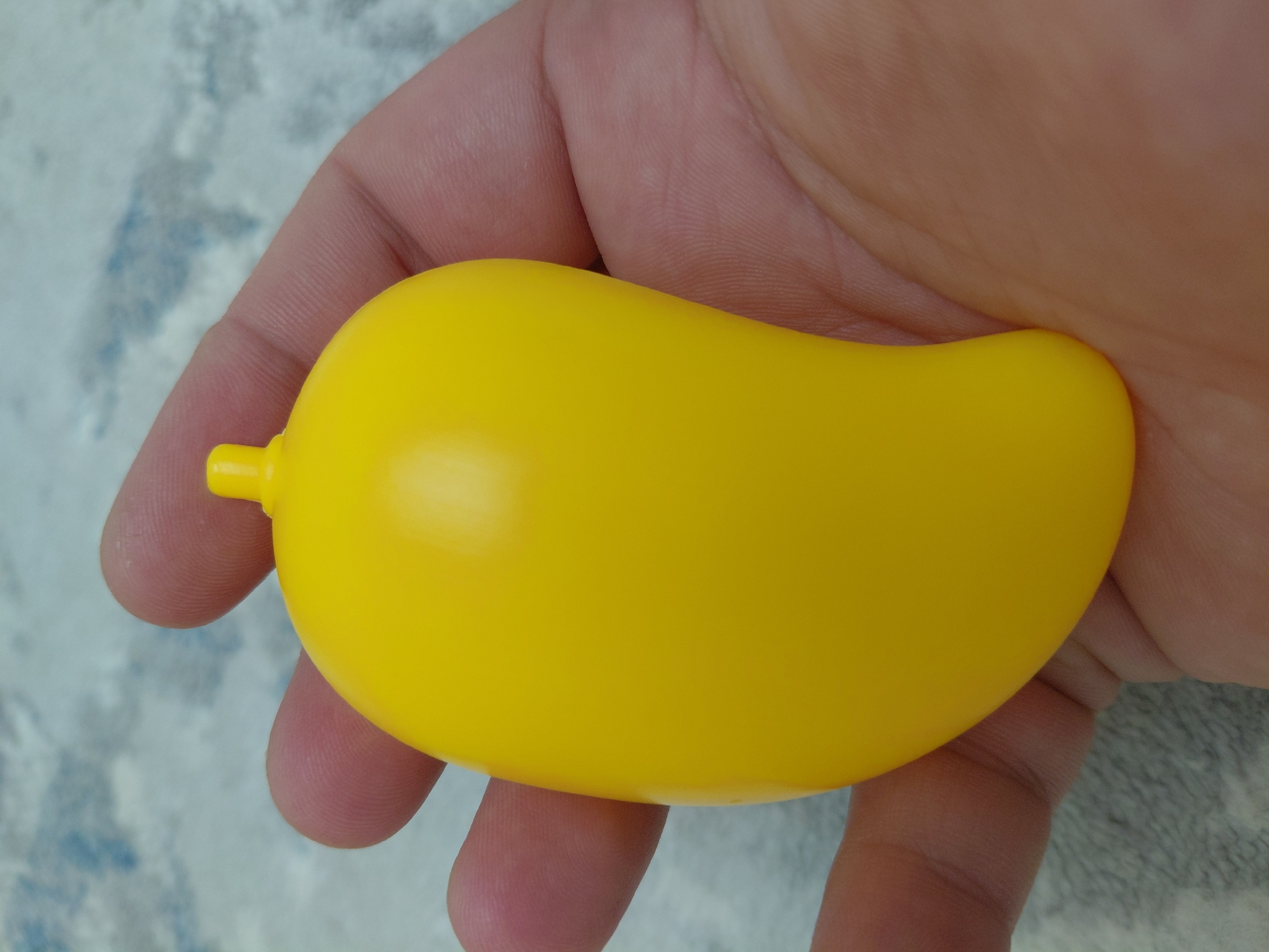 A close-up, overhead shot shows a bright yellow toy mango held in a person's left hand, against a grey, textured background that appears to be a rug or carpet. The mango is smooth and slightly glossy, with a small stem-like protrusion at the top. The hand's fingers and palm are visible, supporting the toy. The background is out of focus, with subtle blue and grey tones. The image does not contain any identifiable landmarks or context to suggest a location in Aktau, Kazakhstan.