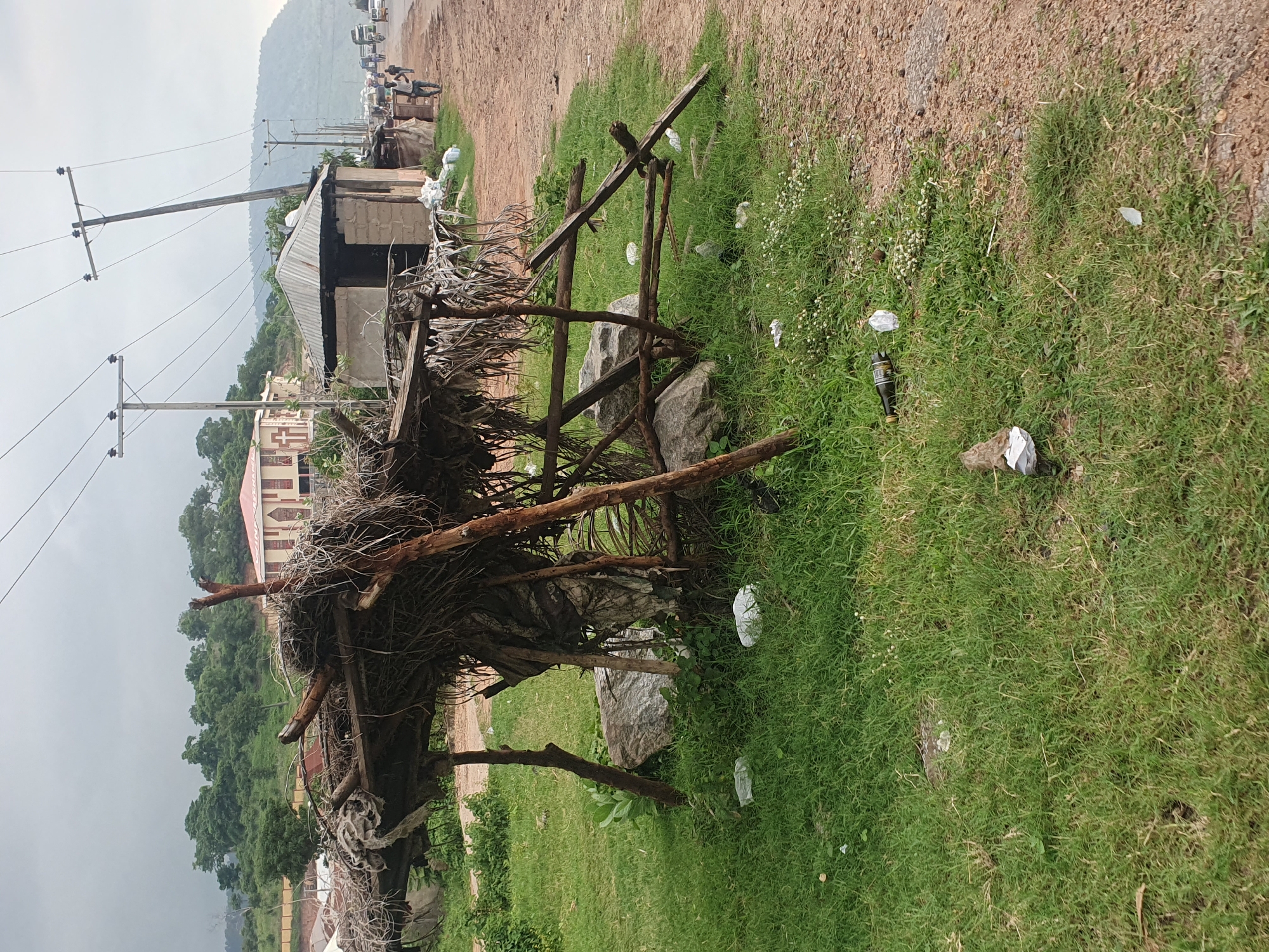 An overcast day in Lokoja, Nigeria, as depicted by the image. In the foreground, a makeshift shelter constructed from branches and dried vegetation stands precariously on four wooden posts. Several large rocks are visible beneath the shelter. The ground surrounding the shelter is covered in green grass, with scattered pieces of litter, including plastic bags and a glass bottle. To the right of the shelter, a dirt road runs alongside the grassy area. Beyond the road, a cluster of small buildings with corrugated metal roofs is visible, along with two electricity poles topped with insulators. In the middle ground, a larger building with a red roof and light-colored walls can be seen, with a sign that reads "CHAPEL" above the entrance. Further in the background, a lush green hillside rises, dotted with trees and more buildings. The road stretches into the distance, with a line of vehicles, including trucks and cars, suggesting moderate traffic. A few people are walking along the side of the road. The sky is a uniform grey, indicating cloudy weather.