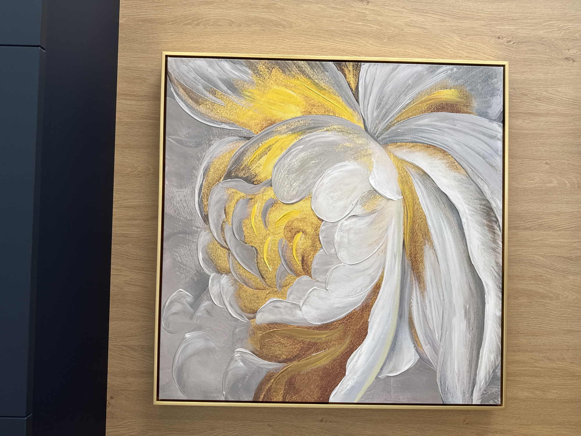 A framed painting hangs on a light-colored, wood-paneled wall. The painting depicts a stylized flower, rendered in shades of grey, white, yellow, and brown, with a gold frame. The flower's petals appear to have a textured, three-dimensional effect. A dark blue object is partially visible on the left side of the frame. The image shows no indications of being in Sofia, Bulgaria.