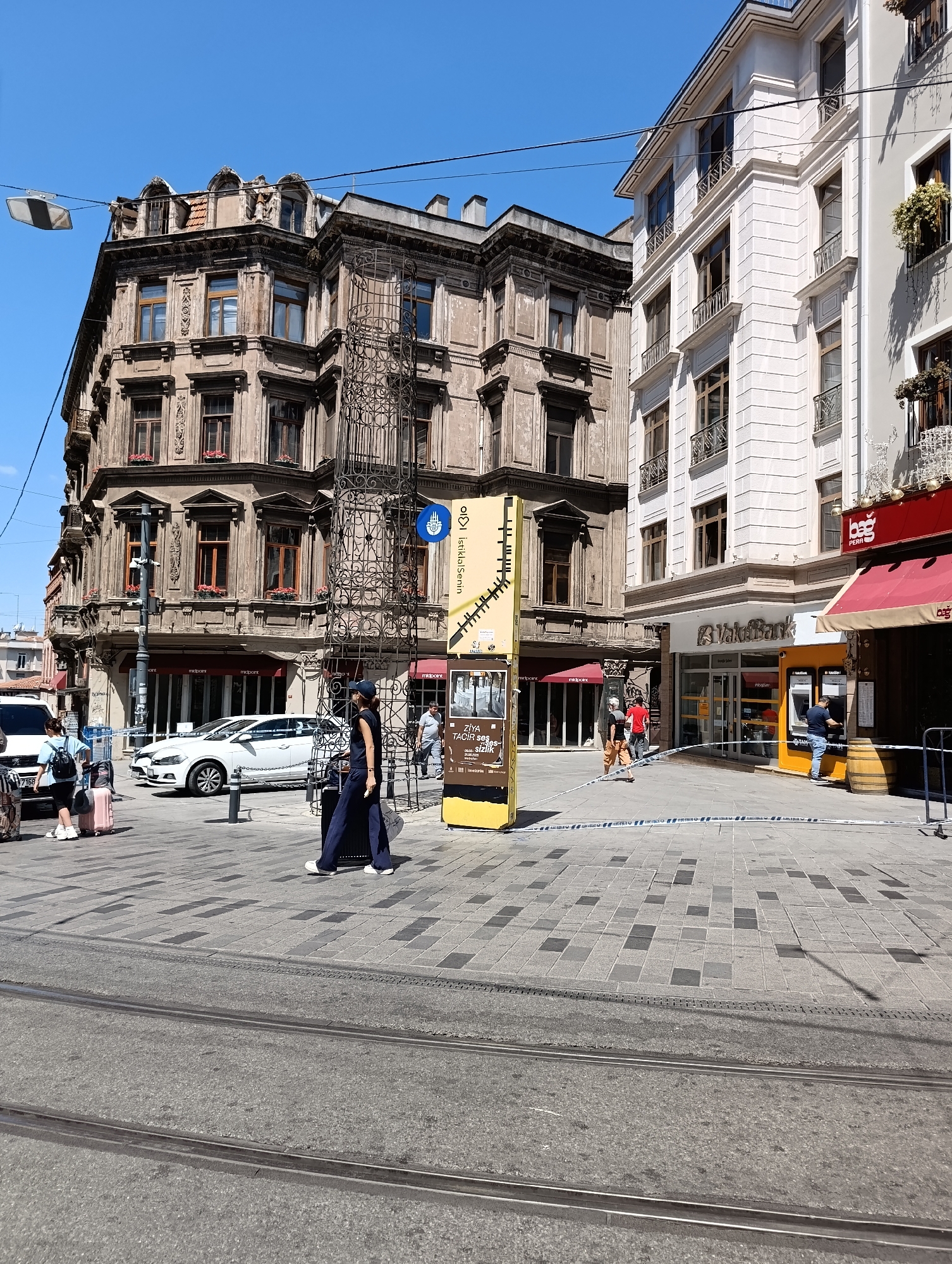 The image depicts a street scene in Beyoğlu, Turkey, under a clear blue sky. The foreground shows tram tracks embedded in a paved pedestrian area. On the right, a white tape barrier is visible, indicating a restricted zone. In the background, there are two multi-story buildings. The building on the left is older, with a weathered facade, ornate architectural details, and several windows. A large, dark metal structure resembling scaffolding or a decorative grill stands in front of it. The building on the right is newer, with a white facade and more modern windows. A sign with the logo "VakıfBank" is mounted on this building, and a person is seen interacting with an ATM outside.
Several people are present in the scene. A woman in a dark outfit is walking with a suitcase. Another woman with a backpack and a pink suitcase is standing on the left side of the image. Two men in casual attire are walking in the middle ground. The overall impression is a busy urban environment on a sunny day, with some areas cordoned off.