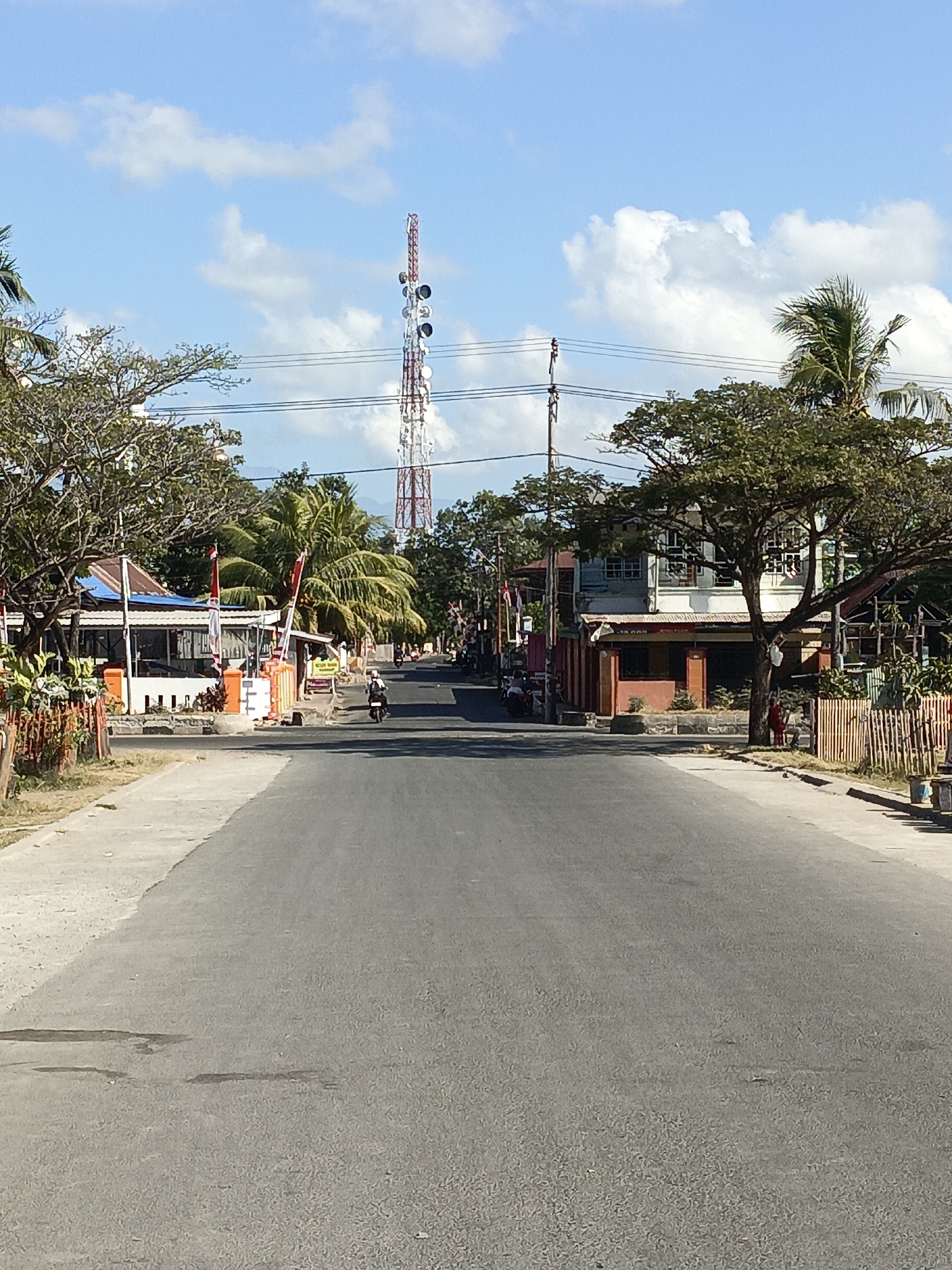 The image shows a road in Bulukumba, Indonesia, on a sunny day. A tall, red and white telecommunications tower stands in the distance, dominating the skyline. On either side of the road are buildings, some with red and white flags displayed. Trees, including palm trees, line the street, providing shade. A few people are visible, one on a motorcycle, and another person standing on the sidewalk. The road is paved and appears to be in good condition. The sky is blue with scattered white clouds. The scene suggests a typical day in a coastal town in Indonesia, with a mix of infrastructure and natural elements.
