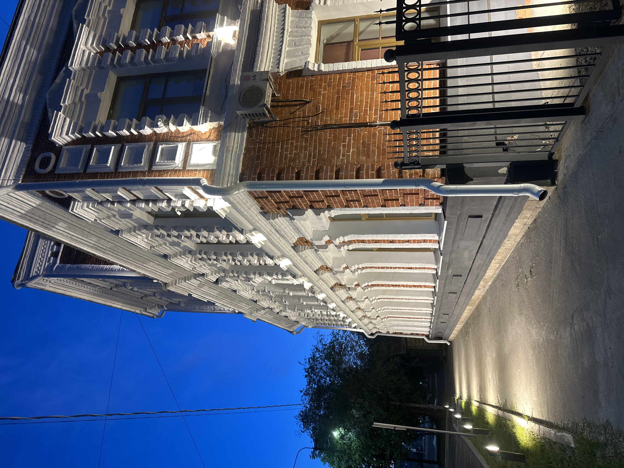 The image captures a dimly lit, narrow pathway bordered by a historic brick building on the right and trees on the left. The building, characterized by intricate white stucco detailing and red brickwork, is illuminated by strategically placed lights, casting a warm glow on its facade. A black metal fence runs along the side of the building, separating it from the pathway. The pathway itself is lit by a series of modern, low-lying lamps, creating a welcoming ambiance. The sky above is a deep twilight blue, indicating that the photograph was taken at dusk or in the early evening.
The building appears to be situated on a street in Blagoveshchensk, Russia, possibly a historical district, given its architectural style. The scene is devoid of any people or overt activity, presenting a tranquil and somewhat serene atmosphere. The overall impression is one of quiet evening beauty, highlighting the architectural grandeur of the building and the careful illumination of the surrounding pathway.