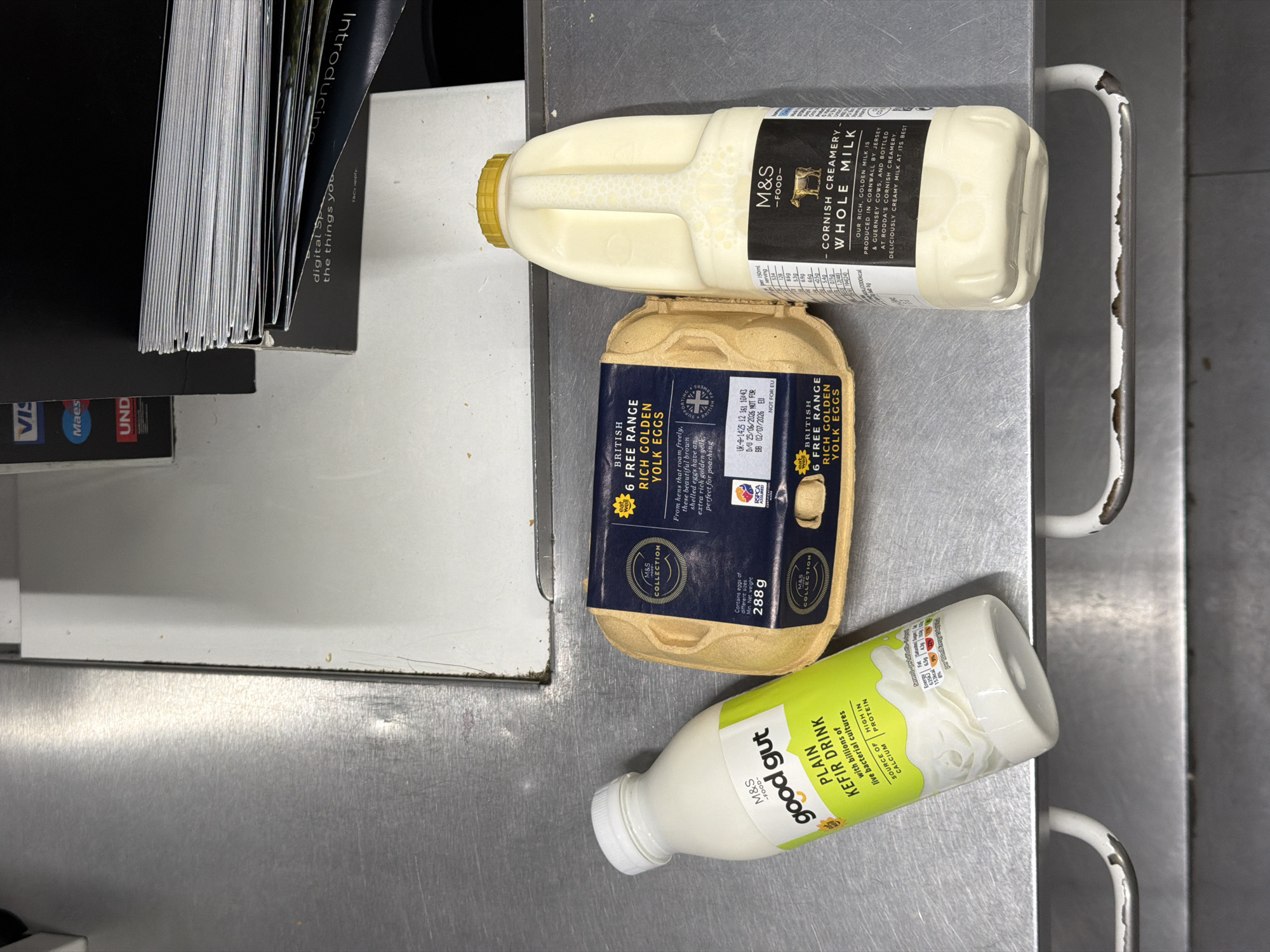 The image shows a retail counter in a supermarket in London, United Kingdom. On the counter are three M&S (Marks & Spencer) branded products: a bottle of "Good Gut Plain Kefir Drink," a carton of 6 British Free Range Rich Golden Yolk Eggs, and a bottle of "Cornish Creamery Whole Milk." The counter surface is metallic and appears to be part of a checkout or display area. In the background, a stack of brochures or magazines is visible, along with payment card terminals. The scene is well-lit and appears to be a standard retail environment.