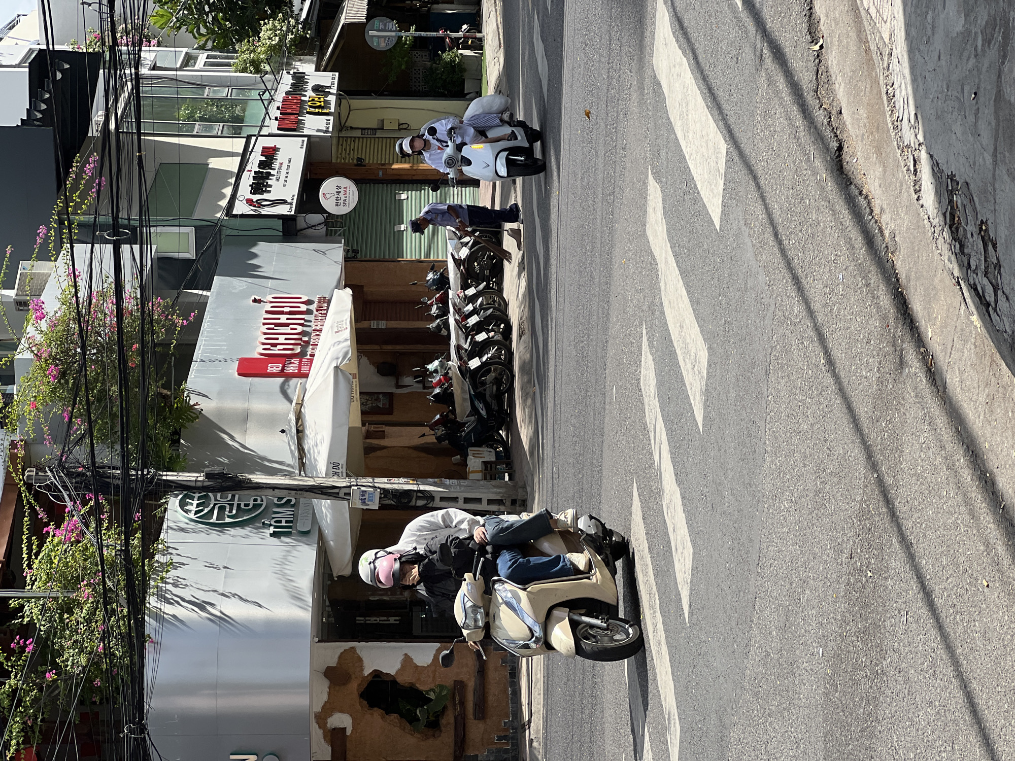 The image captures a street scene in Nha Trang, Vietnam, on a sunny day. In the foreground, a pedestrian crossing is visible on the asphalt road. Two people are riding on a light-colored scooter, with the driver wearing a white helmet and a gray hoodie, and the passenger behind wearing a pink helmet and a dark jacket. The scooter is in motion, crossing the pedestrian walkway. Further down the street, a white scooter is approaching, ridden by a person wearing a white helmet and a white top. To the left, a row of parked motorcycles is lined up along the sidewalk. A street cleaner wearing a blue uniform and a cap is sweeping the sidewalk with a broom. The buildings lining the street feature modern architecture with glass windows and signage. Lush green trees with pink flowers are visible on the upper levels of some buildings, with numerous black electrical wires crisscrossing overhead. The overall environment suggests a typical urban setting in Vietnam.