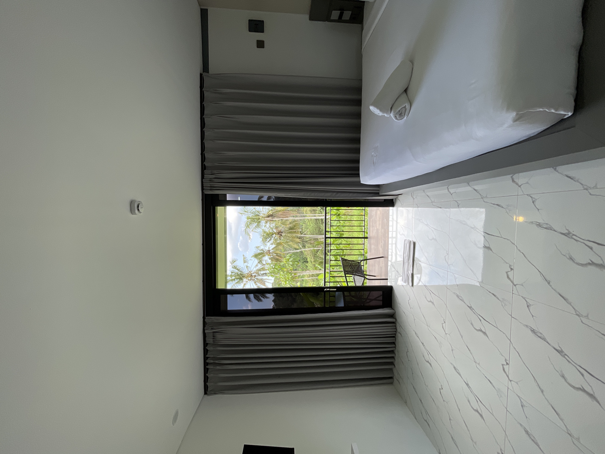 The image shows a hotel room in Penebel, Indonesia, with a large window or sliding glass door opening to a balcony overlooking a lush tropical landscape with palm trees. The room features a white tiled floor with a marble pattern, light gray curtains, and a bed with white linens and rolled-up white towels. A white rug is placed on the floor near the balcony entrance. The balcony has a black railing and a single black chair. A smoke detector is visible on the white ceiling. The overall impression is of a clean and modern hotel room with a focus on the natural view.