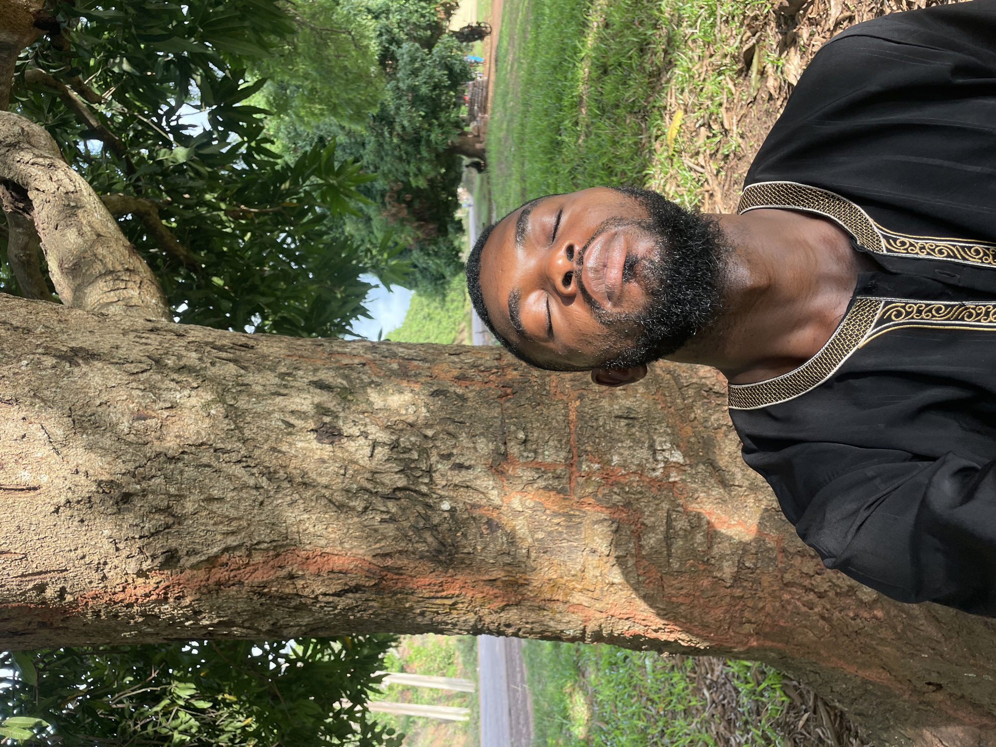 A man with closed eyes leans against a tree in Ogbe, Nigeria. He has dark skin, a beard, and wears a black shirt with gold trim around the collar. The tree has rough, brown bark. In the background, there is green grass and other trees. A road or path is visible beyond the tree trunk.