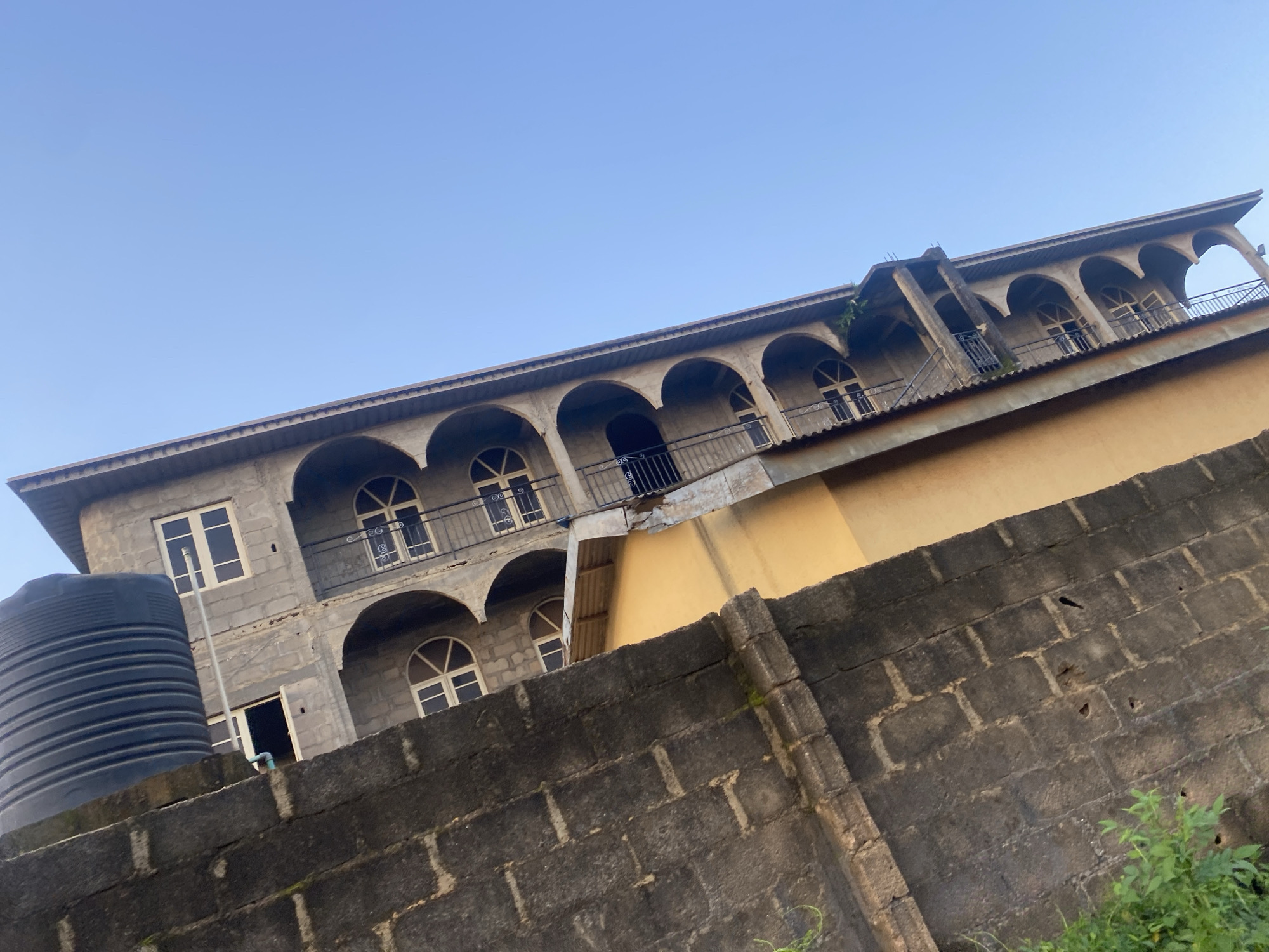 This image captures a low-angle view of an unfinished multi-story building, likely in Ikorodu, Nigeria, under a clear blue sky. The building's facade features a series of arched openings on each floor, some of which contain windows with decorative white frames. The construction appears to be of concrete blocks, with some sections covered in yellow paint. A large black water tank is visible at the top left of the frame, mounted on a section of grey brick wall that occupies the left foreground. Lush green vegetation grows at the base of this brick wall, suggesting an outdoor setting. The scene appears to be devoid of human presence.
