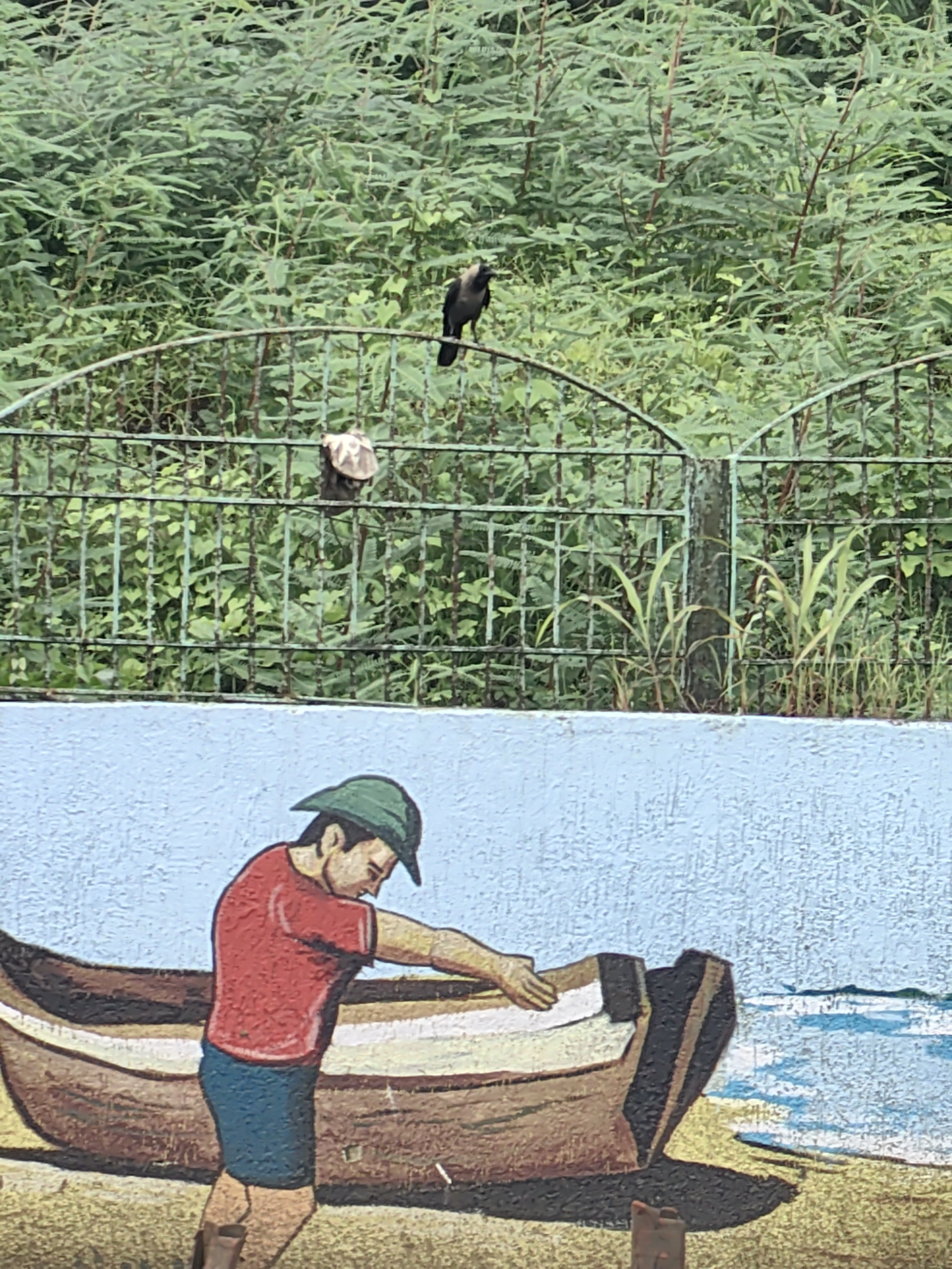 The image shows a mural painted on a wall in Mira-Bhayander, India. The mural depicts a person, likely a young male, wearing a red shirt, blue shorts, and a green hat, working on a boat. The boat is brown and appears to be on a beach with a representation of the ocean in the background. The mural is painted on a textured, light blue wall. Above the mural, a green metal fence is visible, with dense green foliage behind it. Two birds and a turtle are perched on the fence. The lighting suggests it is daytime.