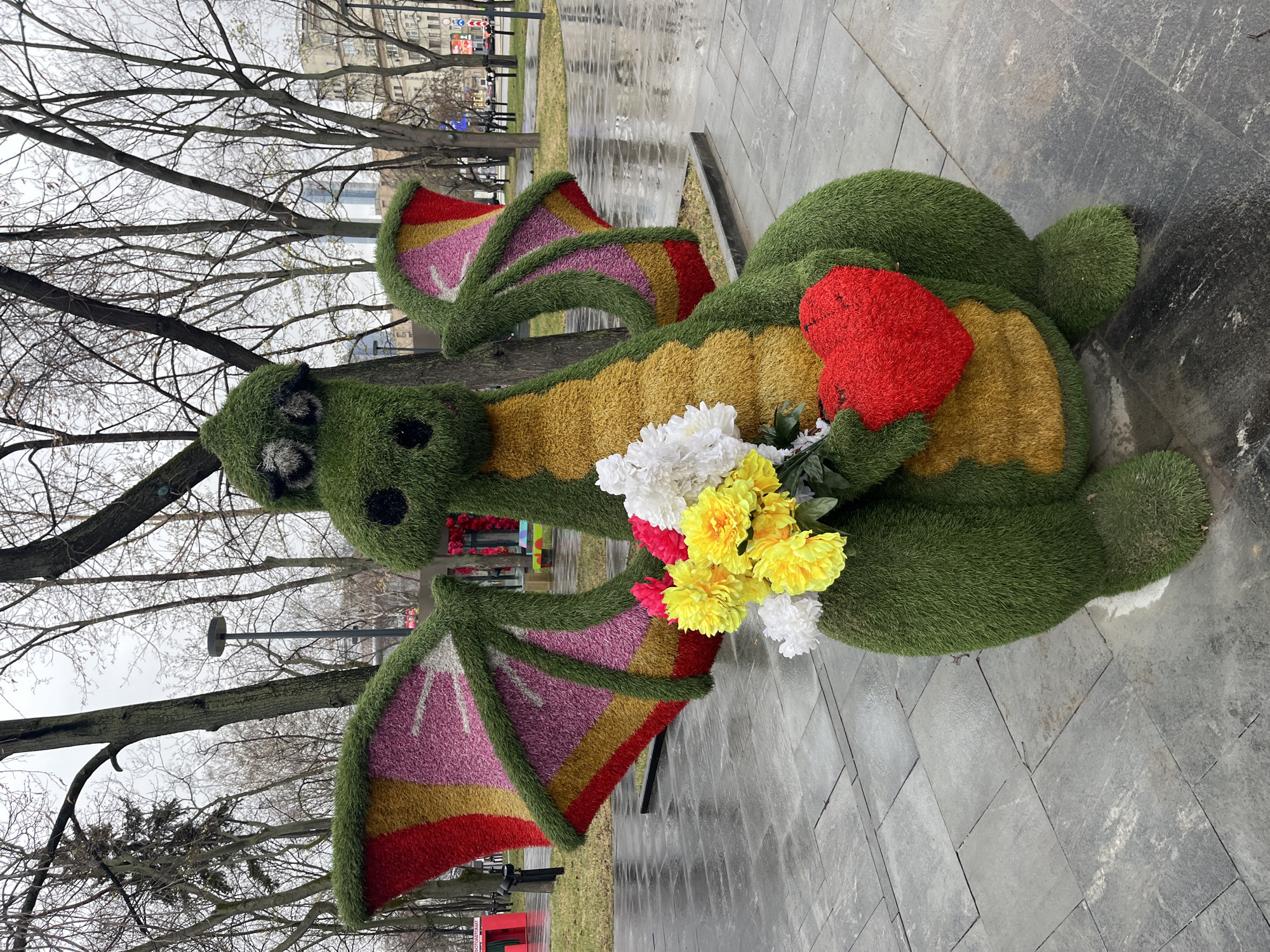 The image shows a topiary dragon figure holding flowers and a heart, located on a paved area. The dragon is made of green grass-like material, with a yellow belly. Its wings have bands of red, yellow, and pink. The flowers are white and yellow with a bit of red. The dragon is also holding a red heart.
The background consists of bare trees and buildings in the distance. The location is likely Moscow, Russia, based on visual cues and the context of the request.