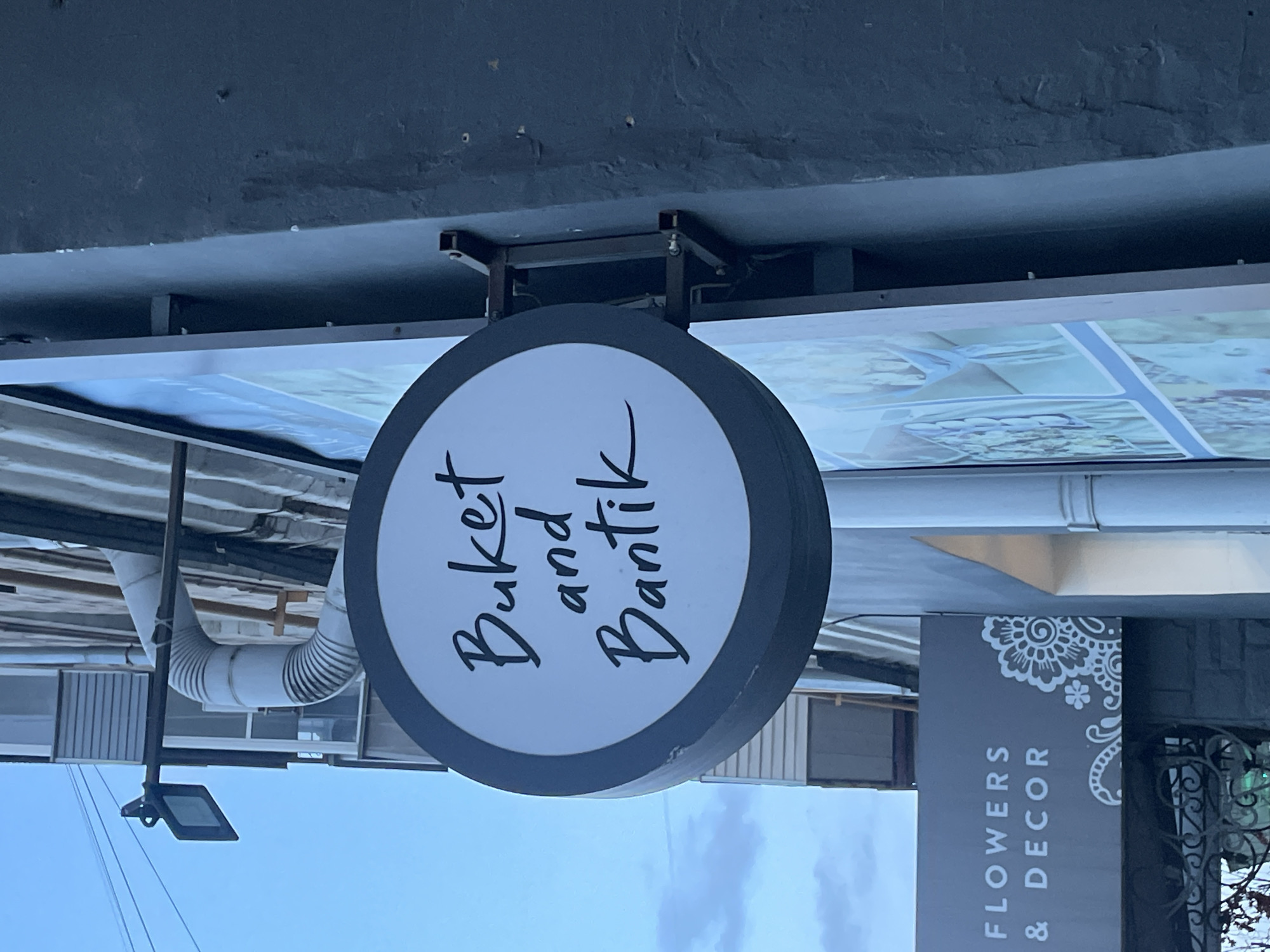 A street-level photograph shows a business sign in Krasnoyarsk, Russia. The primary sign is circular with a white background and a dark gray border, displaying the text "Buket and Bantik" in a script font. The sign is attached to a building via a metal frame. Adjacent to this sign is another sign reading "Flowers & Decor" vertically. An external ventilation duct and a street light fixture are also visible. The sky is light blue with some cloud cover. The photograph is taken from a low angle, suggesting the photographer is standing close to the ground.