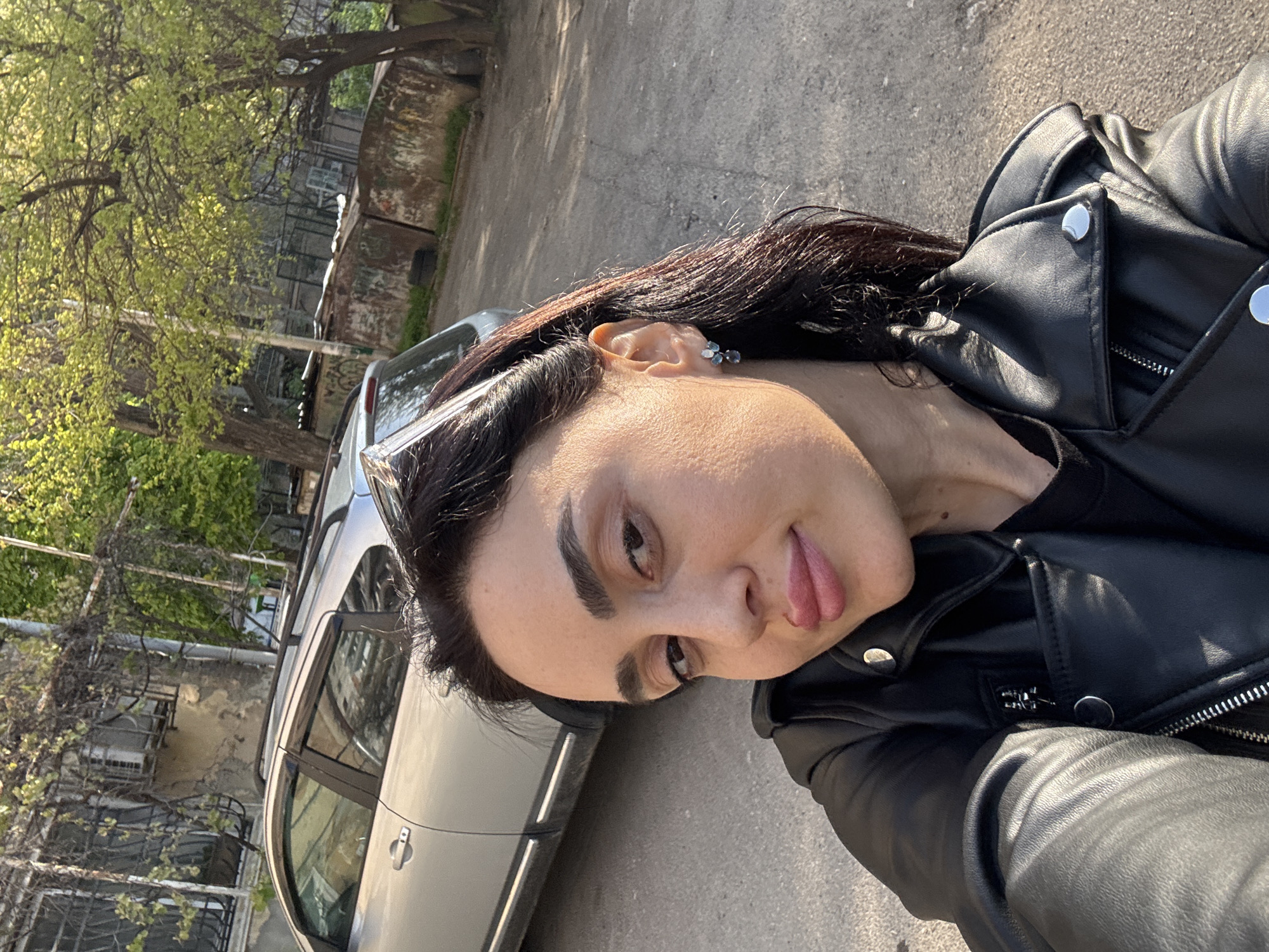 A woman is depicted in a selfie-style shot, sticking her tongue out. She is wearing a black leather jacket. Behind her is a silver car parked on a city street, and in the background, there are trees and buildings. The location is inferred to be Tbilisi, Georgia, based on contextual information (which is not visible in the image).