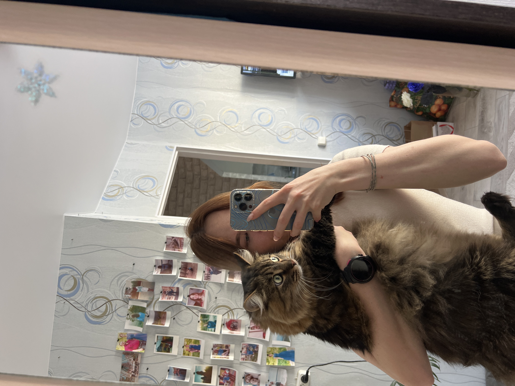 A woman is taking a selfie in a mirror while holding a brown and black tabby cat. The woman is holding a blue iPhone 13 or 14 in her left hand and has a black smartwatch on her left wrist. The room has wallpaper with a pattern of swirling lines and circles, and a collection of small photographs pinned to the wall. There is a doorway visible behind the woman. There is a snowflake decoration on the ceiling and a floral arrangement on a shelf. The image was taken in a room that appears to be located in the city of Chaikovsky, Russia.