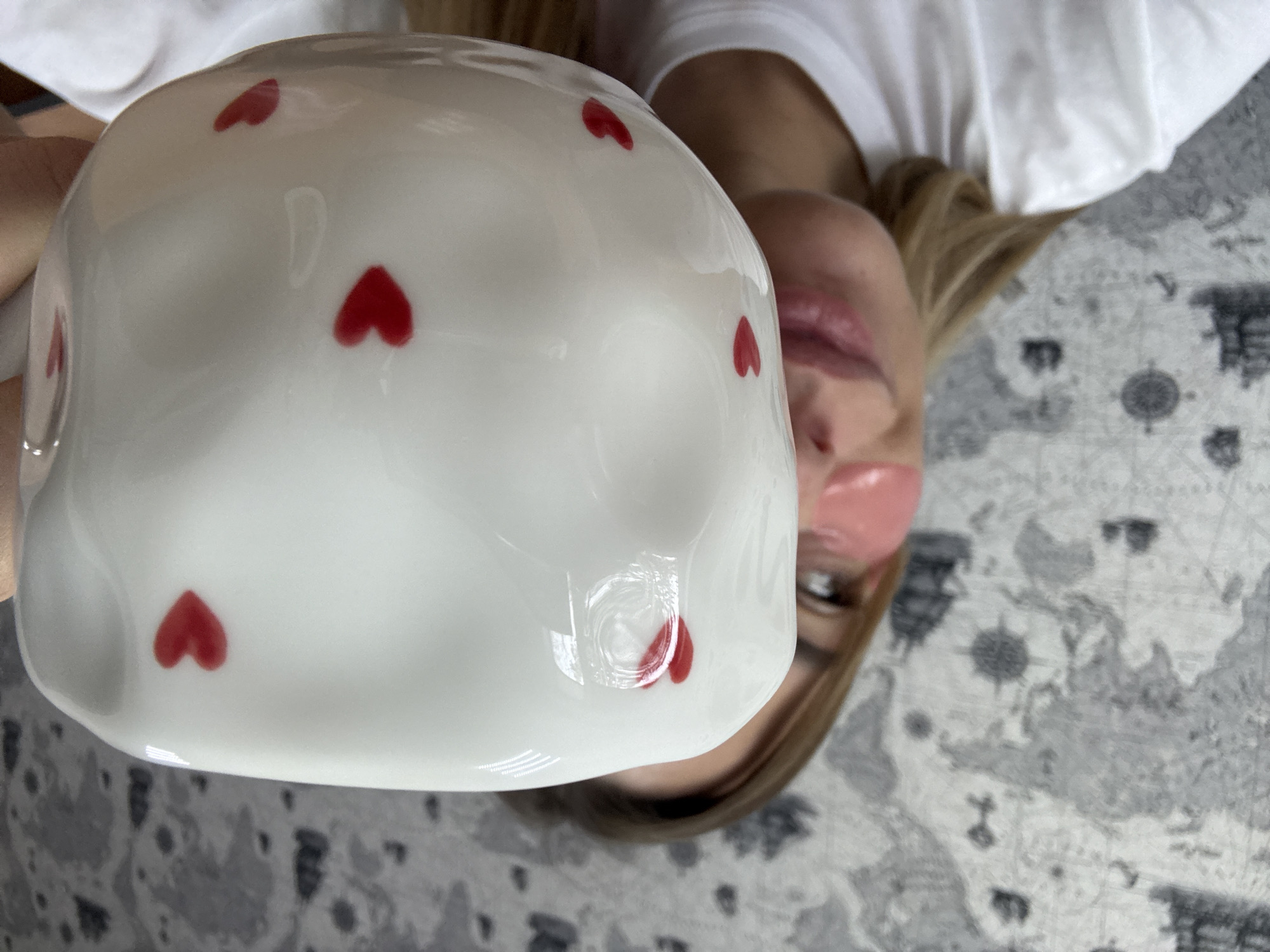 A woman lies on a gray patterned rug while holding a white mug with red heart decorations. She is wearing pink under-eye patches and a white t-shirt. The background includes a partial view of the rug and her blonde hair.